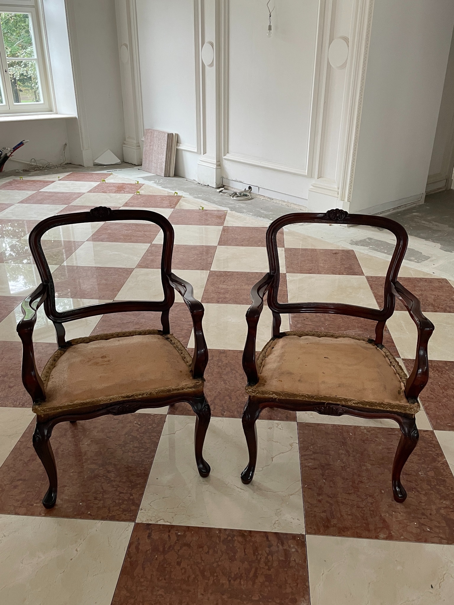 Dwa krzesła w stylu retro na podłodze w szachownicę, wnętrze w trakcie remontu, widoczne listwy przypodłogowe i okno. Krzesła z ciemnego drewna z beżowymi siedziskami.