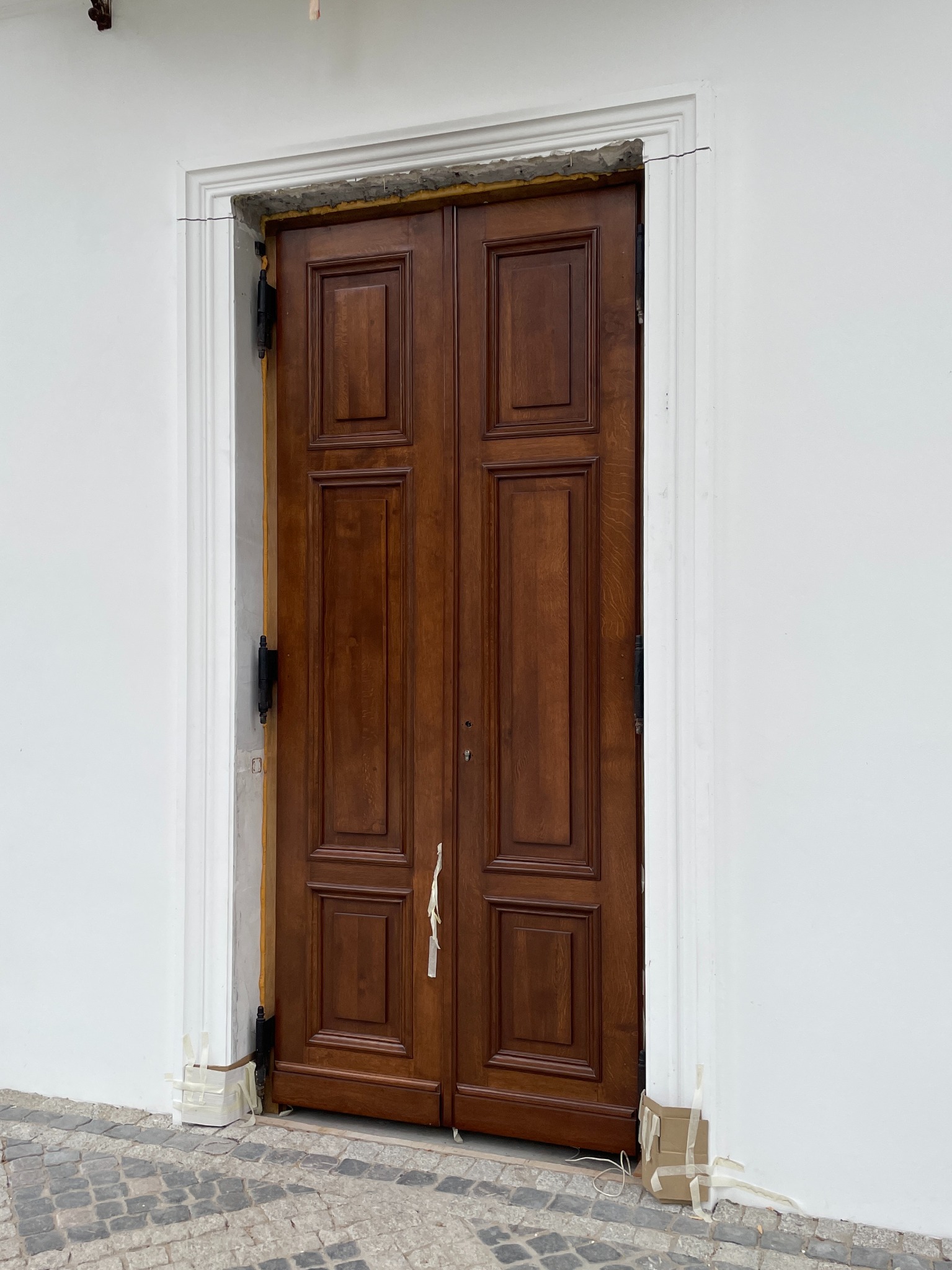 Montaż dwuskrzydłowych drzwi z ciemnego drewna w białej futrynie. Widoczne zabezpieczenia taśmą, na zewnątrz budynku, kostka brukowa.