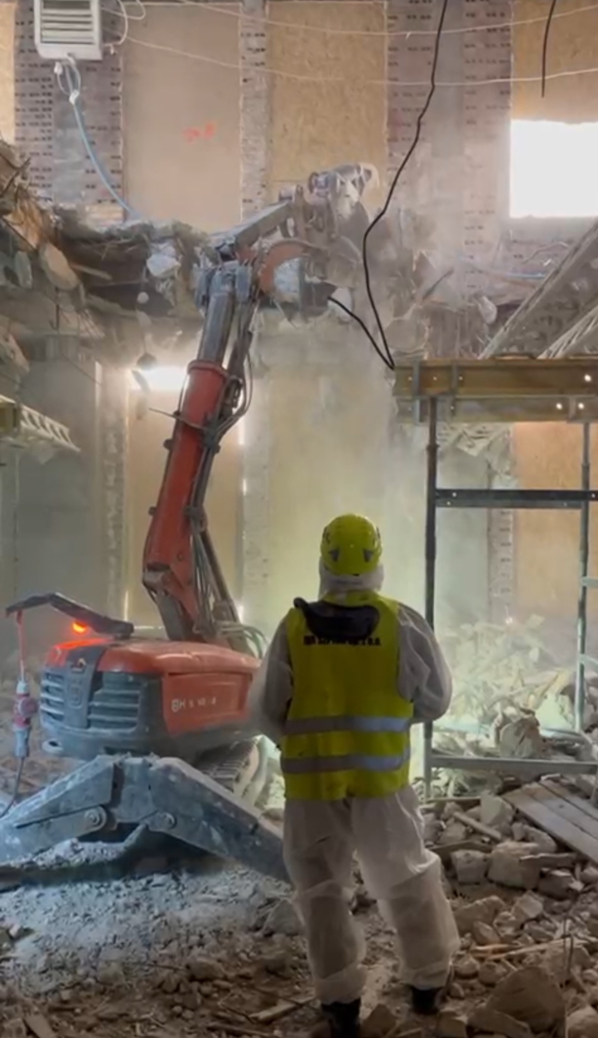 Pracownik w kombinezonie nadzoruje pracę koparki burzącej ścianę budynku. Gruz i pył w powietrzu, widoczne elementy konstrukcji.