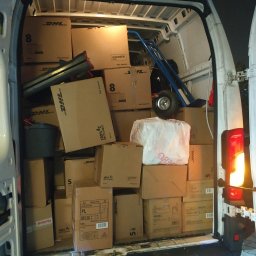 BIKIN'BRAIN SPÓŁKA Z OGRANICZONĄ ODPOWIEDZIALNOŚCIĄ - Wnętrze busa wypełnione kartonami DHL, w tle wózek transportowy. Widok z tyłu pojazdu, światło stopu włączone. Nocna sceneria, transport towarów.