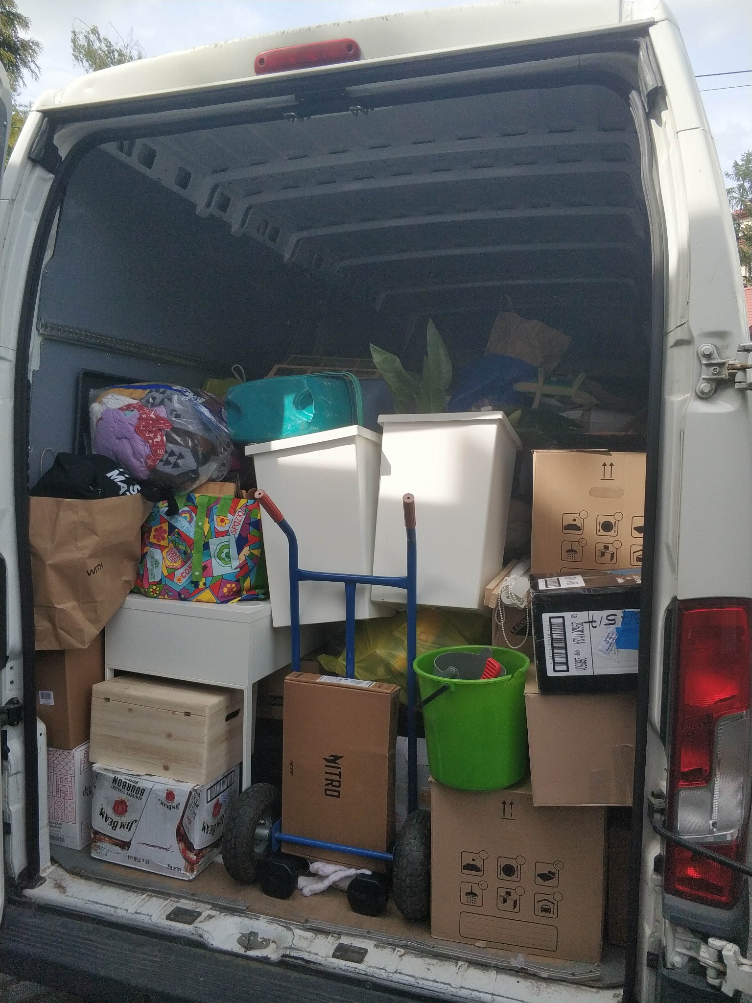 Wnętrze białego busa wypełnione kartonami, torbami i wiadrami, przygotowanego do transportu. W tle widoczne rośliny doniczkowe i inne przedmioty.