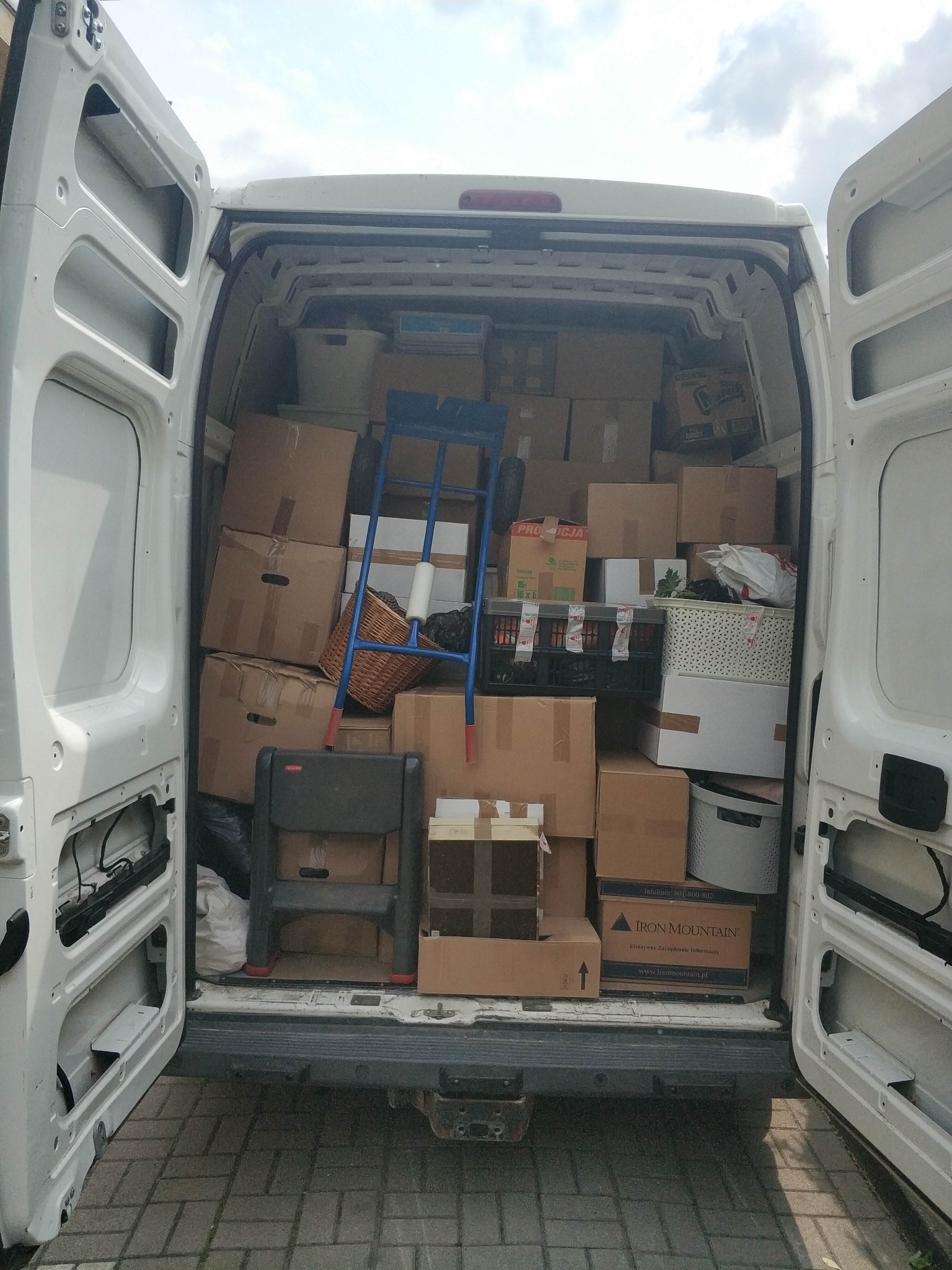 Wnętrze białego busa wypełnione kartonami, wózkiem transportowym i schodkami. Otwarte drzwi pojazdu, widoczna kostka brukowa na zewnątrz.