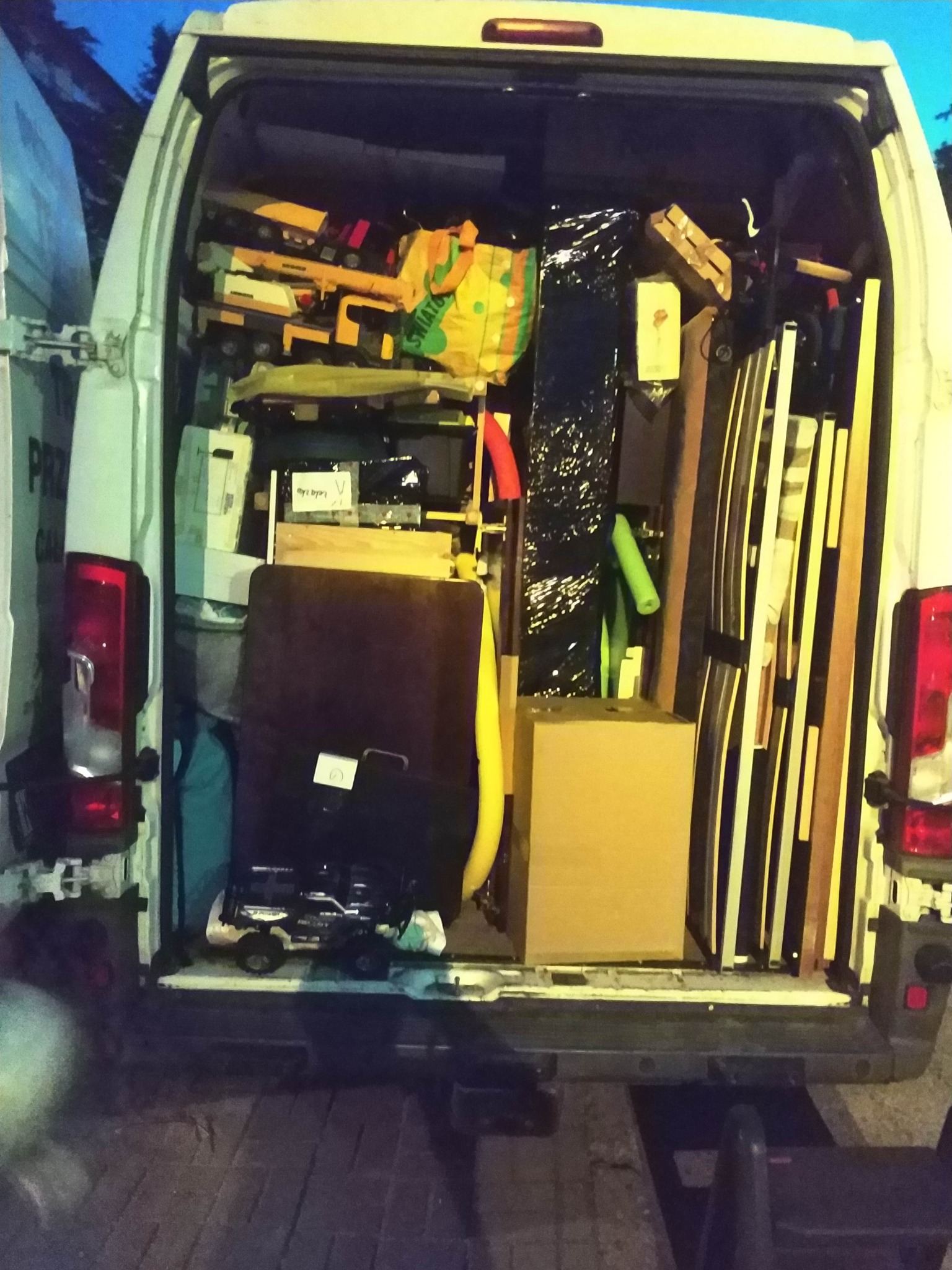 Otwarty bagażnik białego busa wypełnionego różnorodnymi przedmiotami, w tym zabawkami, meblami i zapakowanymi rzeczami, gotowymi do transportu.