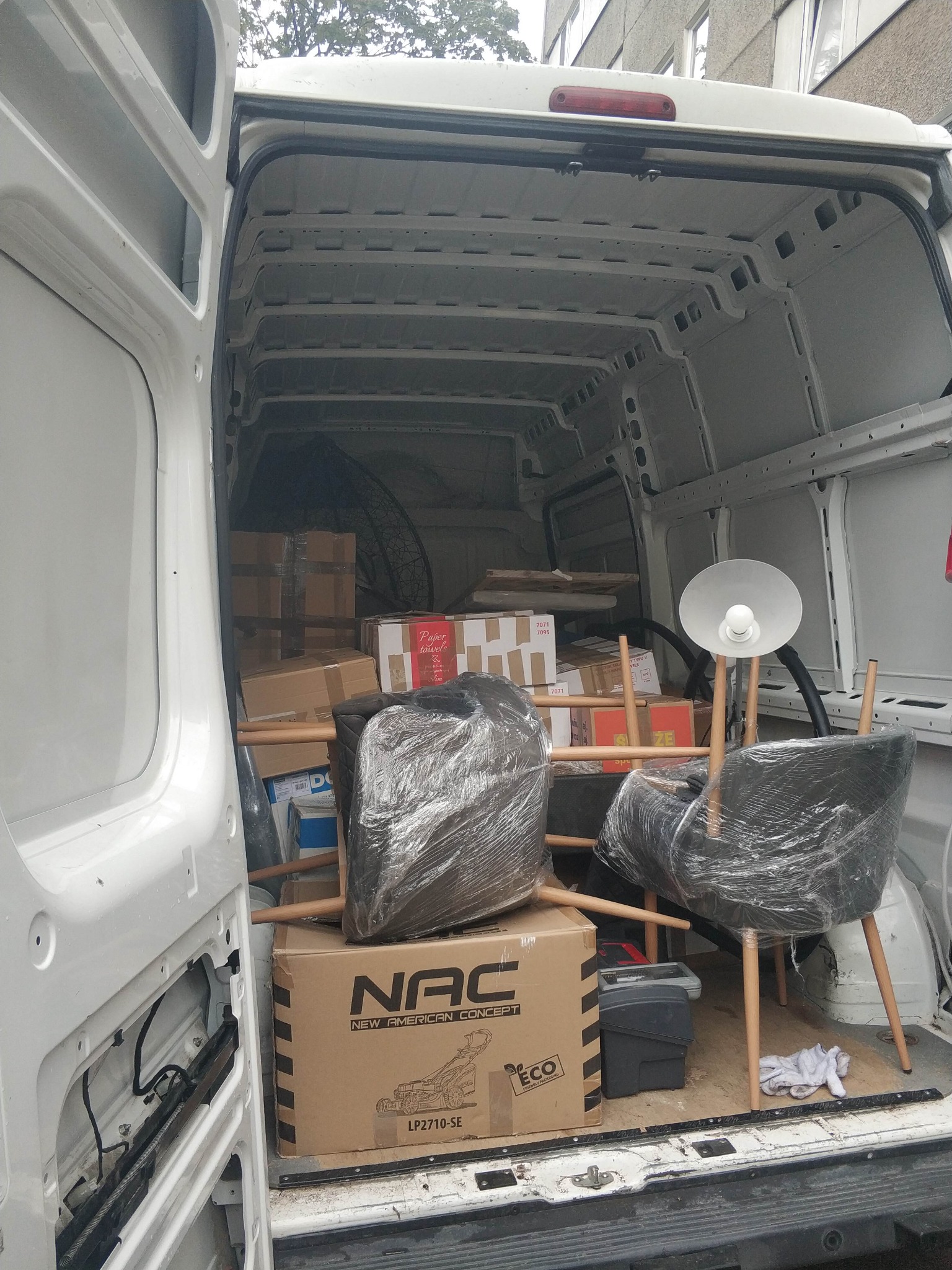 Wnętrze białego busa wypełnione zapakowanymi meblami, kartonami i lampą. Widoczne krzesła owinięte folią, skrzynka narzędziowa i karton z napisem NAC.