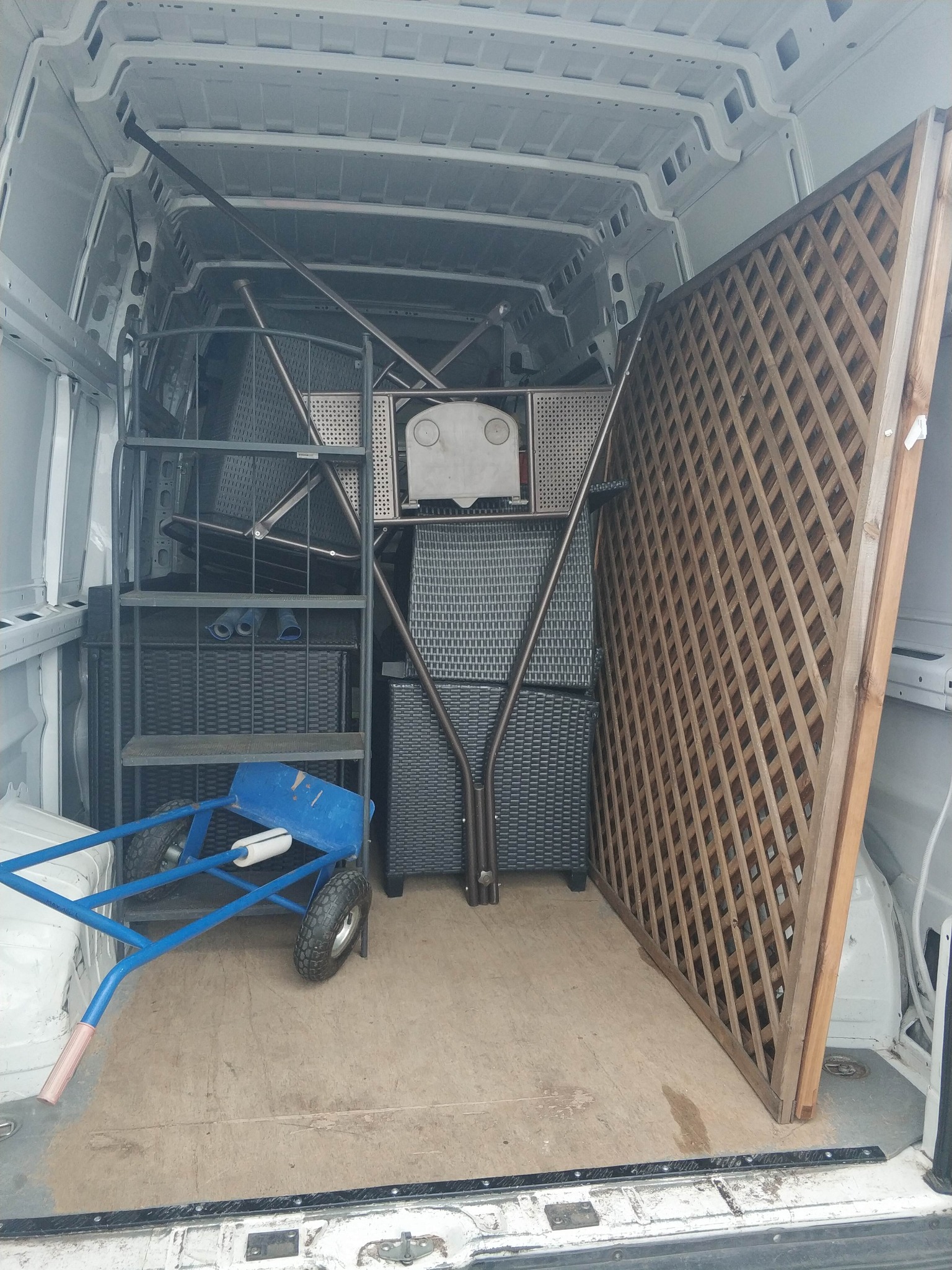 Wnętrze busa wypełnione meblami ogrodowymi (krzesła, stoliki) i drewnianym parawanem. Niebieski wózek transportowy w tle.