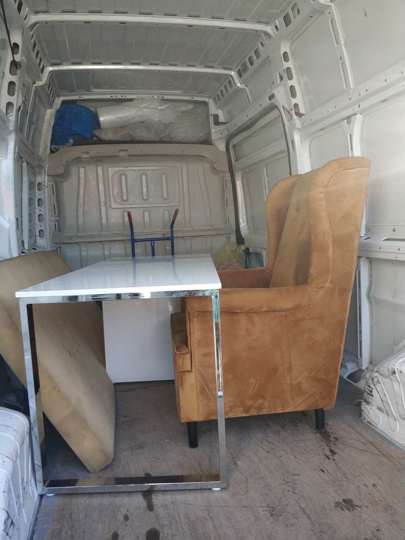 Wnętrze białego busa z meblami: fotel uszak w kolorze musztardowym i stół z białym blatem na chromowanych nogach. Widoczne zabezpieczone przedmioty.