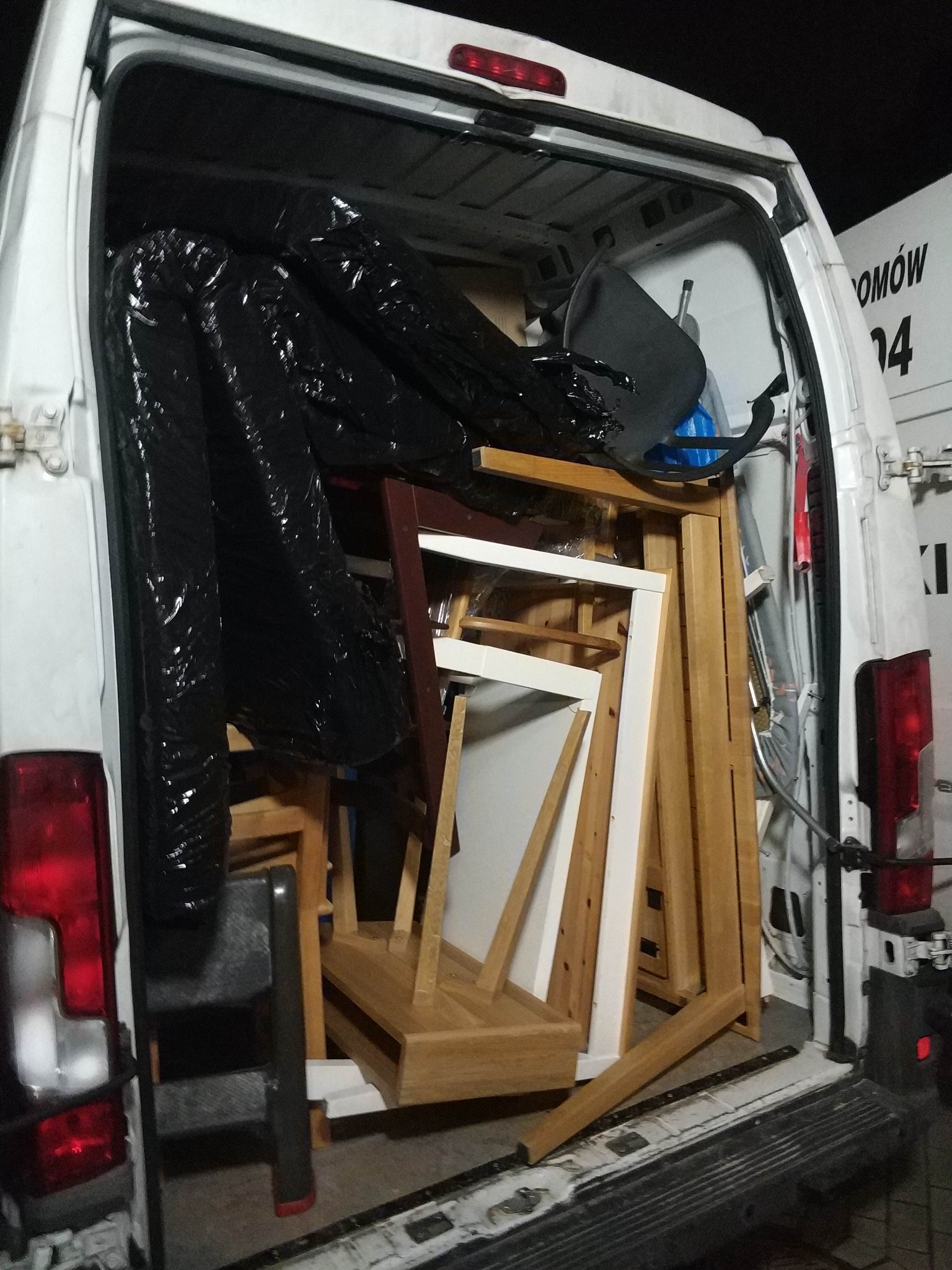 Wnętrze białego busa wypełnione meblami, w tym krzesłami i stołami, zabezpieczonymi folią, gotowymi do transportu. Widok z tyłu pojazdu.