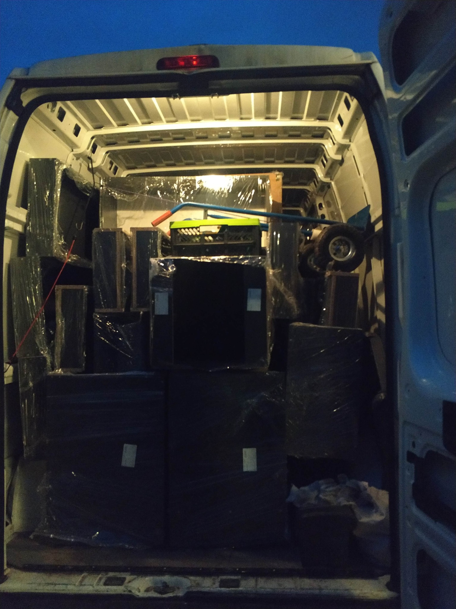 Wnętrze białego busa wypełnione po brzegi zapakowanymi w folię meblami i sprzętami podczas przeprowadzki, widok z tyłu pojazdu, wieczorne oświetlenie.