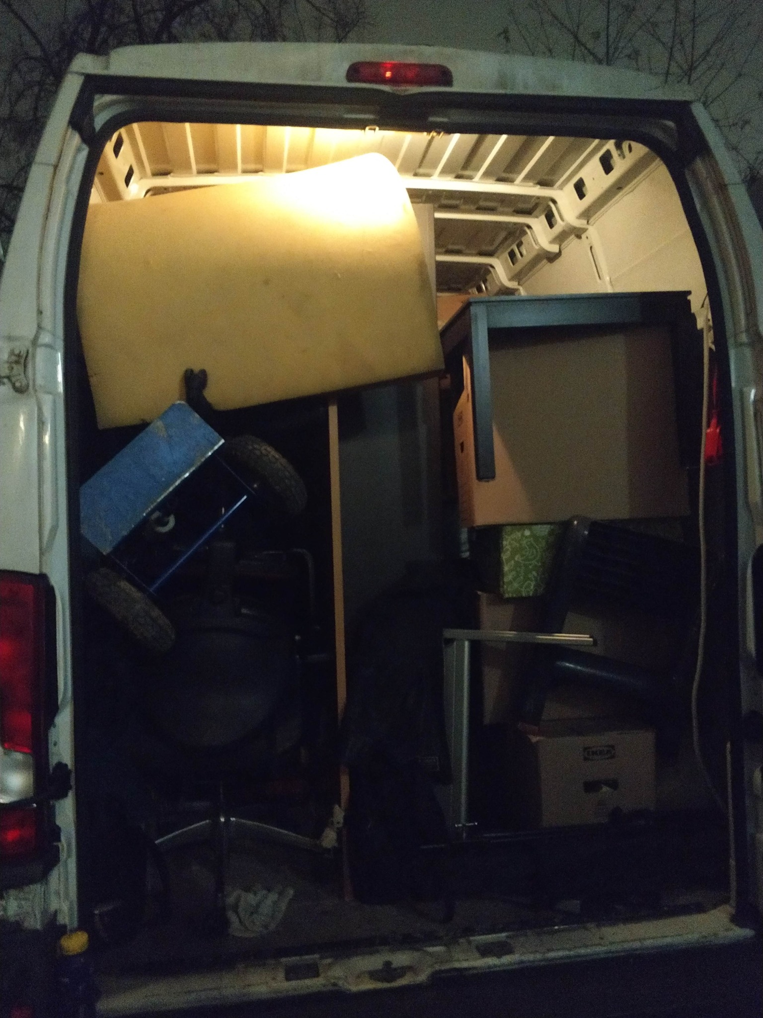 Załadunek mebli i kartonów do wnętrza białego busa, widok z tyłu. Wnętrze oświetlone, widoczny materac, stół i pudełko IKEA. Nocna sceneria.