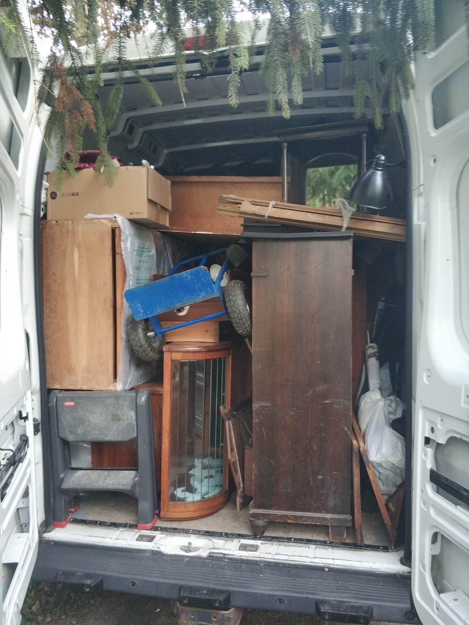 Wnętrze białego busa wypełnione meblami, kartonami i wózkiem transportowym. Widoczne elementy wyposażenia wnętrz, sugerujące przeprowadzkę. Gałązki drzewa w górnej części kadru.