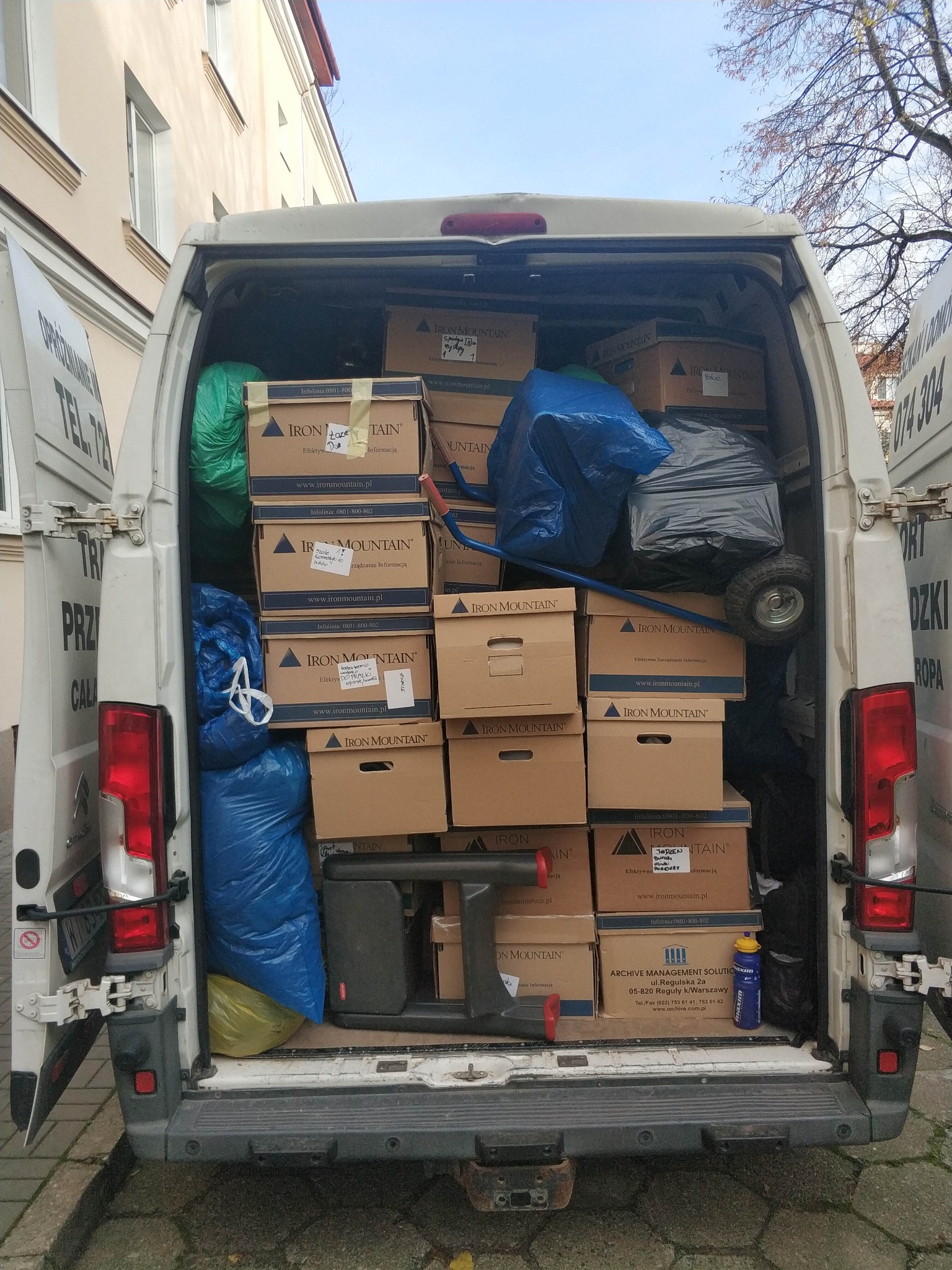 Wnętrze białego busa wypełnione kartonami Iron Mountain i workami, gotowe do transportu. Widoczne detale: wózek transportowy i butelka z napojem.