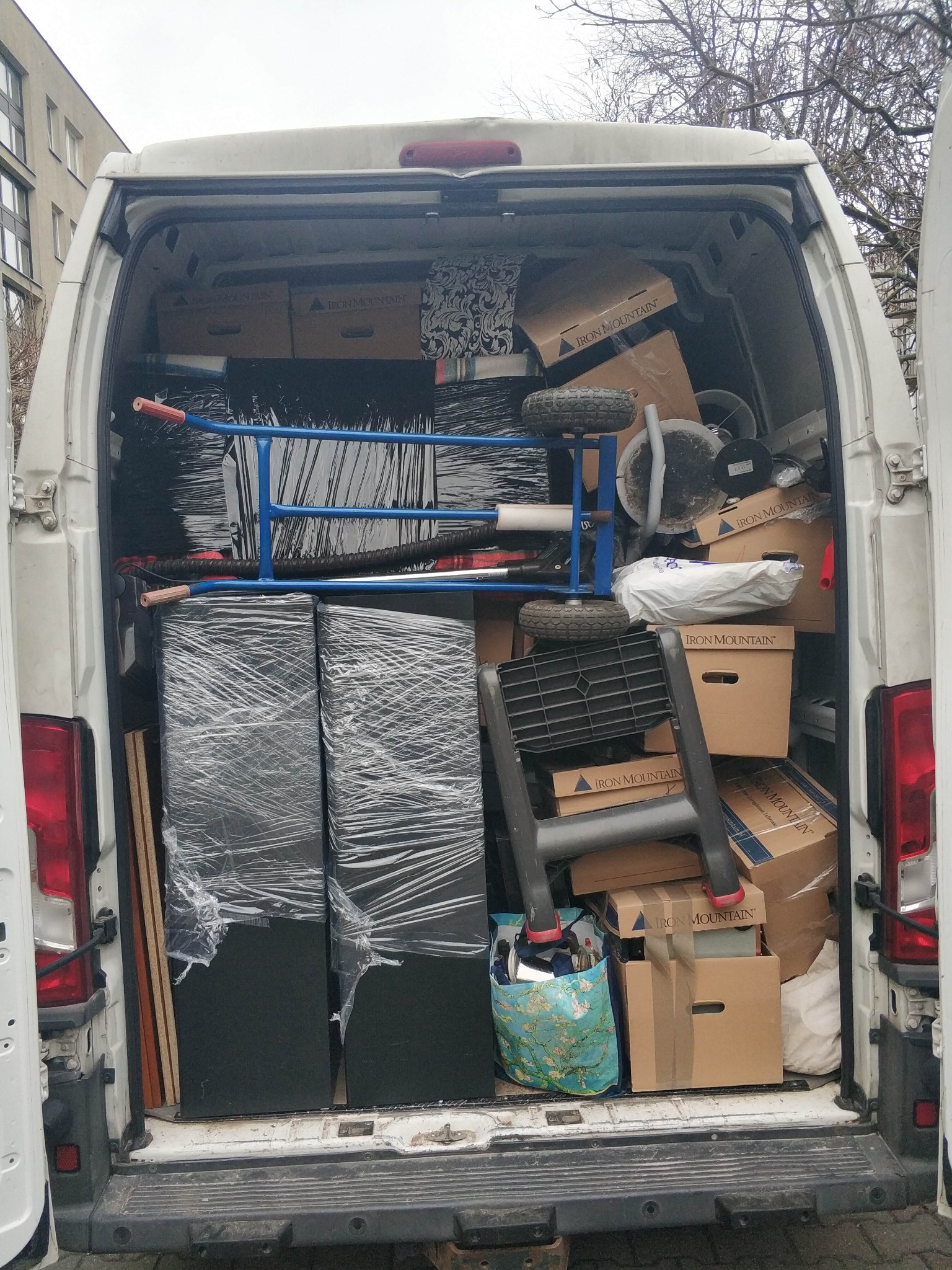 Wnętrze białego busa wypełnione kartonami, meblami owiniętymi folią, wózkiem transportowym i drabiną, przygotowane do transportu. Widok z tyłu pojazdu.