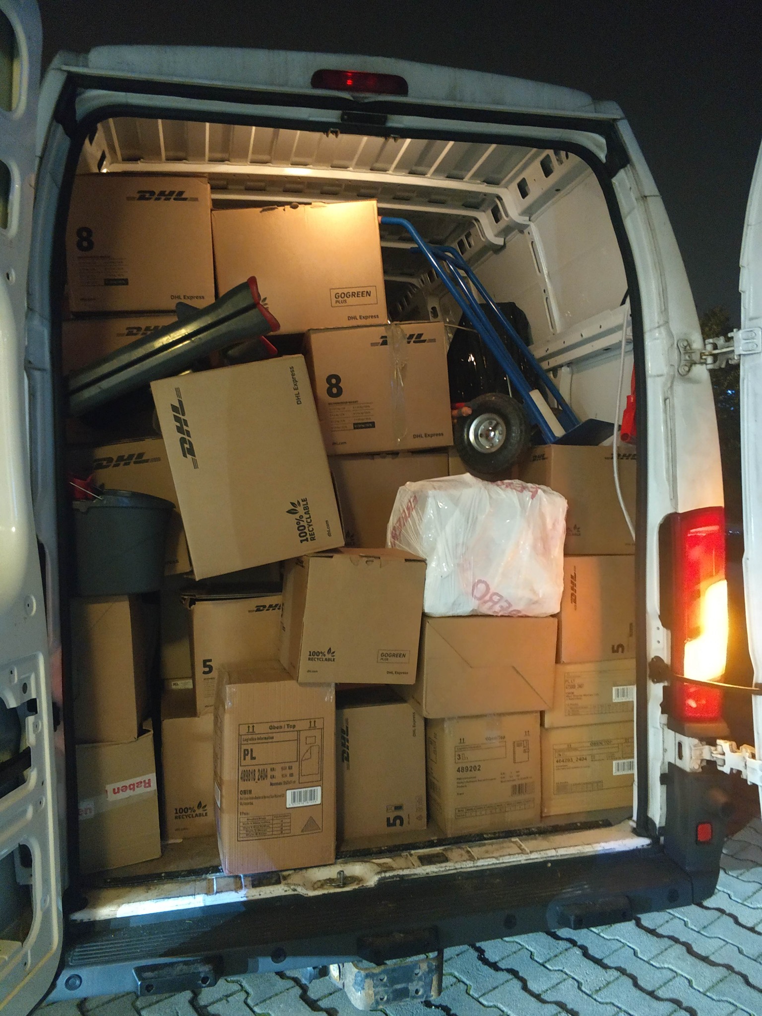 Wnętrze busa wypełnione kartonami DHL, w tle wózek transportowy. Widok z tyłu pojazdu, światło stopu włączone. Nocna sceneria, transport towarów.