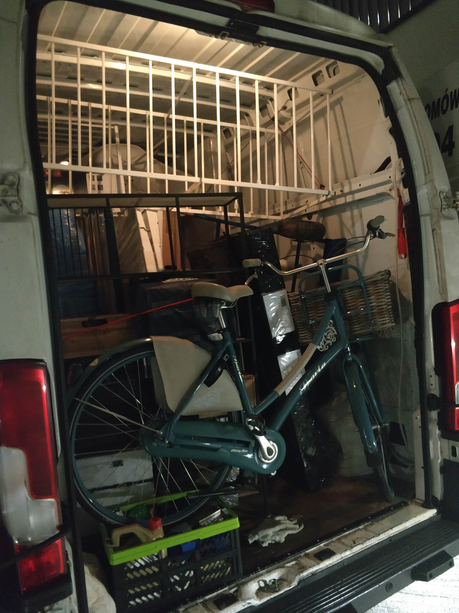 Wnętrze busa załadowane meblami i rowerem miejskim z wiklinowym koszykiem, zabezpieczone kratą. Widok z tyłu pojazdu, oświetlenie wewnętrzne.