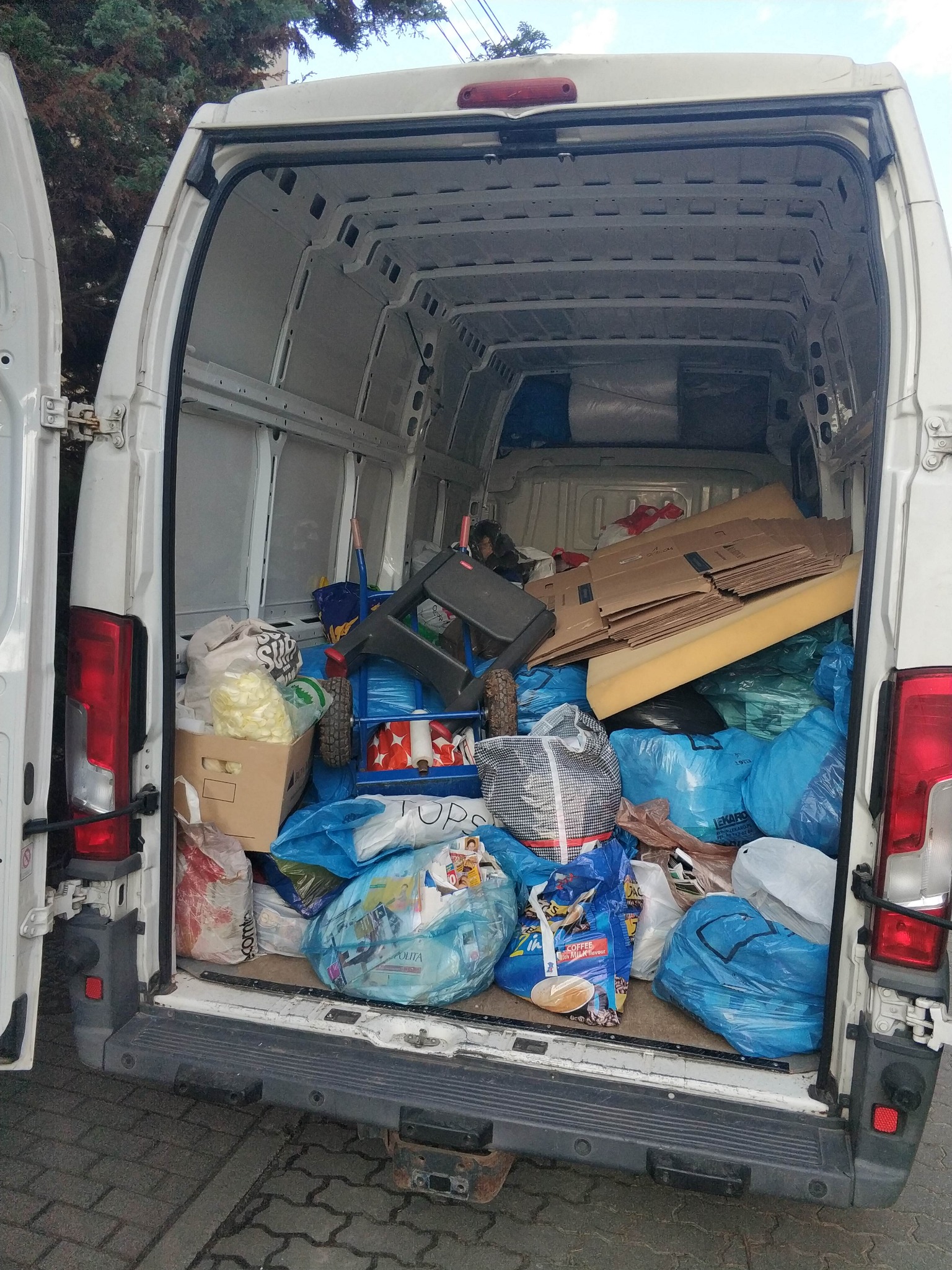 Wnętrze białego busa wypełnione po brzegi kartonami, workami i meblami gotowymi do transportu. Widoczne otwarte tylne drzwi pojazdu.