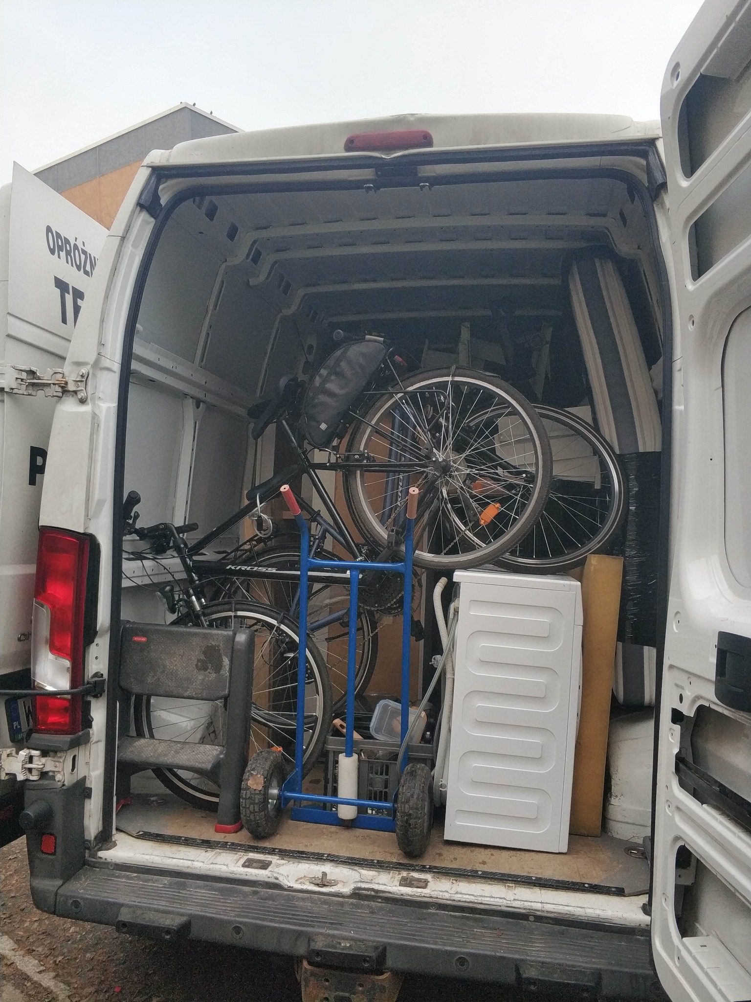 Wnętrze białego busa załadowane rowerami, pralką i kartonami na wózku transportowym. Widok z tyłu pojazdu podczas załadunku.