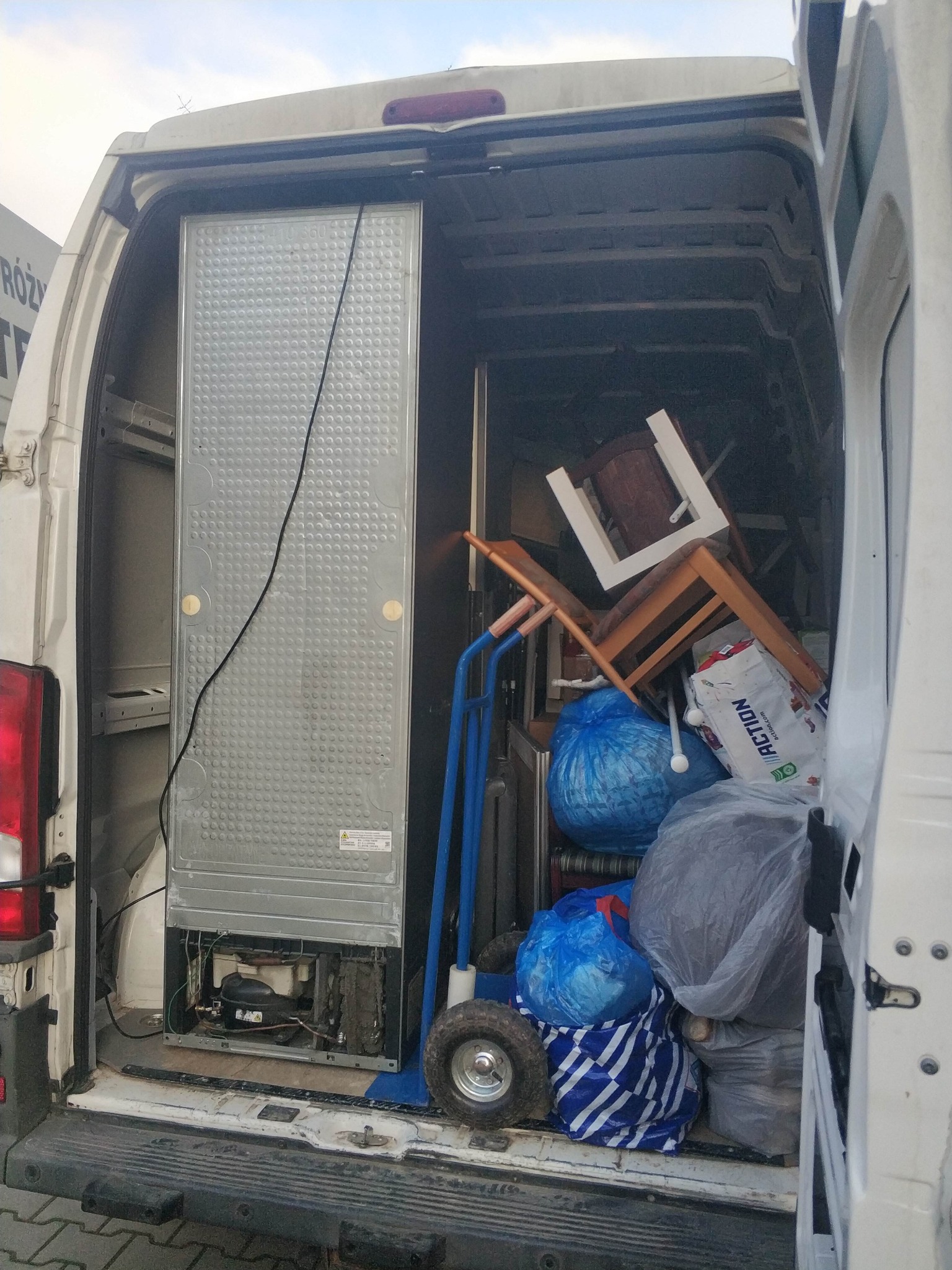 Wnętrze busa załadowane meblami, lodówką i workami podczas przeprowadzki. Widoczny wózek transportowy. Ujęcie z tyłu pojazdu, otwarte drzwi.