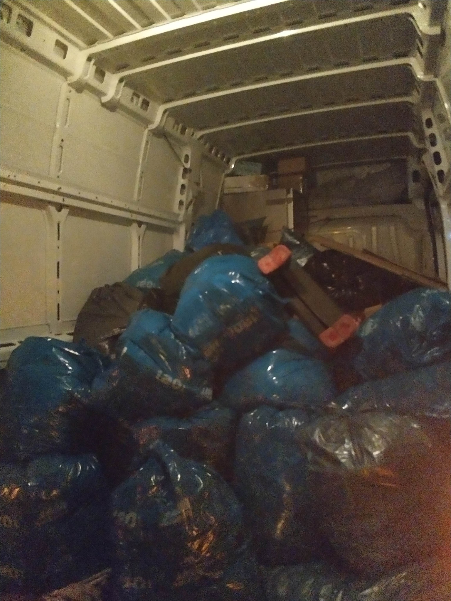 Wnętrze busa wypełnione workami z zawartością, prawdopodobnie podczas przeprowadzki. Widoczne kartony i elementy wyposażenia. Oświetlenie wewnętrzne.
