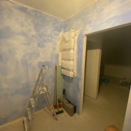 Patryk_Pawluczuk - usługi remontowo budowlane - Pokój w trakcie malowania ścian na niebiesko, drabina, grzejnik owinięty folią, narzędzia malarskie. Widoczny fragment osoby w dolnej części kadru.