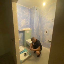 Patryk_Pawluczuk - usługi remontowo budowlane - Remont łazienki: mężczyzna instaluje toaletę w pomieszczeniu z pomalowanymi na niebiesko ścianami. Widoczne narzędzia i materiały budowlane.