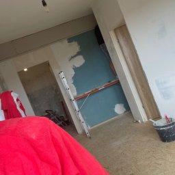 Patryk_Pawluczuk - usługi remontowo budowlane - Wnętrze w trakcie remontu: ściana z częściowo nałożoną gładzią, drabina oparta o ścianę, widoczny fragment podłogi i wiadro z zaprawą. W tle widoczne kolejne pomieszczenie.