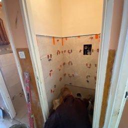 Patryk_Pawluczuk - usługi remontowo budowlane - Układanie płytek w małej łazience z użyciem poziomicy i krzyżyków dystansowych. Widoczny fragment instalacji hydraulicznej. Praca w toku, wnętrze w trakcie remontu.