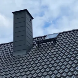 Grafitowy komin z blachy trapezowej i okno dachowe na ciemnej dach&oacute;wce karpi&oacute;wce, na tle błękitnego nieba z chmurami. Widok z dołu.