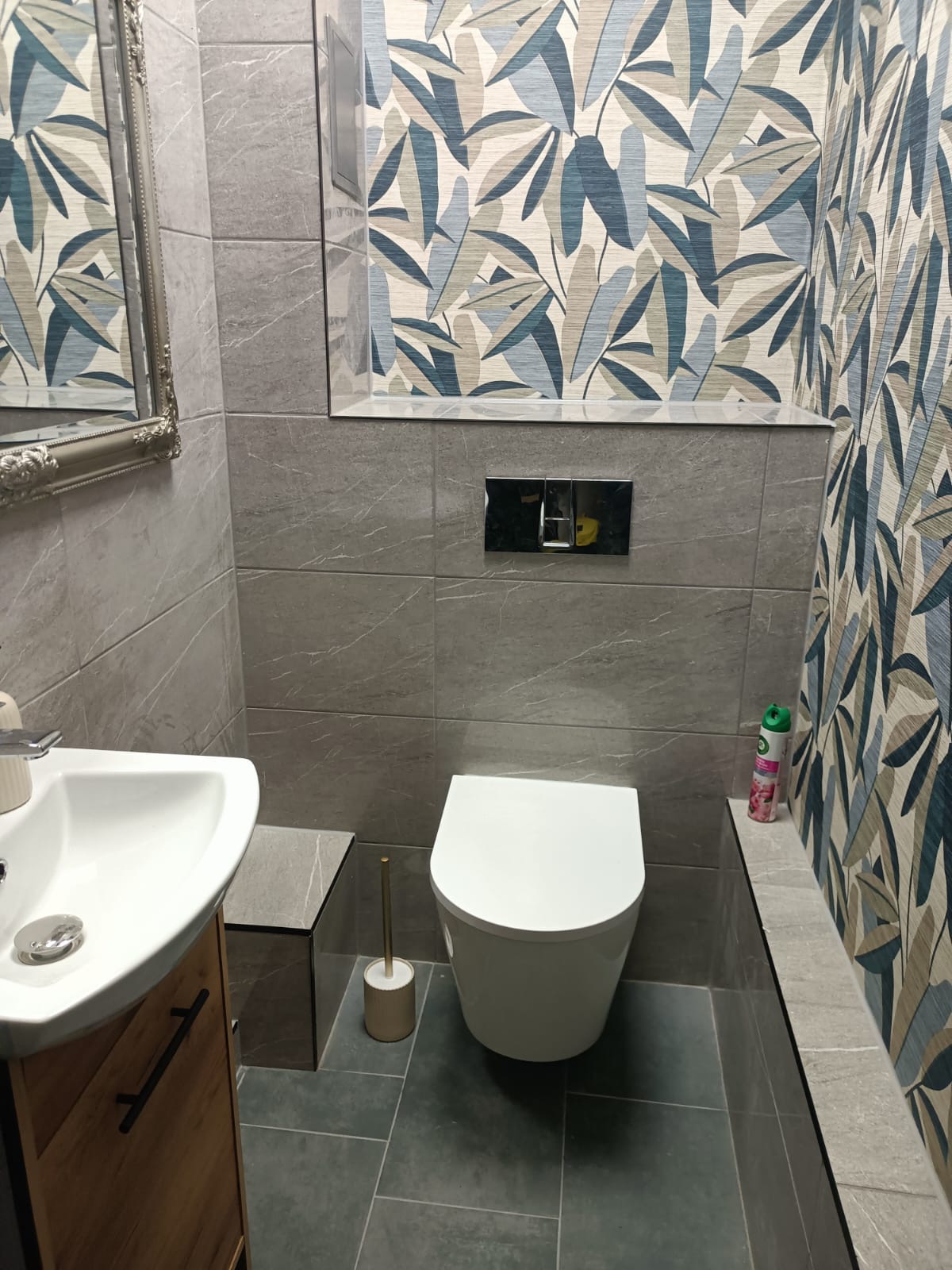 Wykończona łazienka w szarościach z akcentem roślinnym na tapecie. Umywalka z szafką, toaleta i lustro z ozdobną ramą. Widoczne płytki imitujące kamień i nowoczesna armatura.