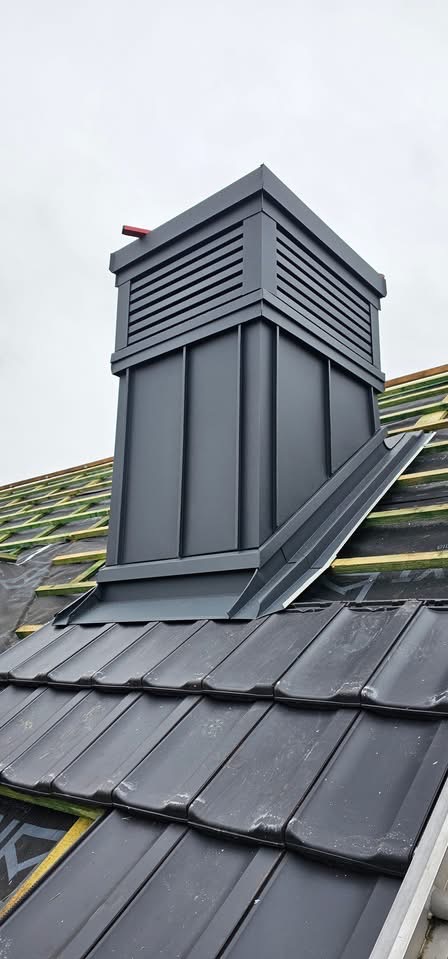 Szary komin wentylacyjny z blachy na dachu z czarnej dachówki ceramicznej. Widoczne elementy konstrukcji dachu. Kąt ujęcia z dołu, pochmurne niebo w tle.