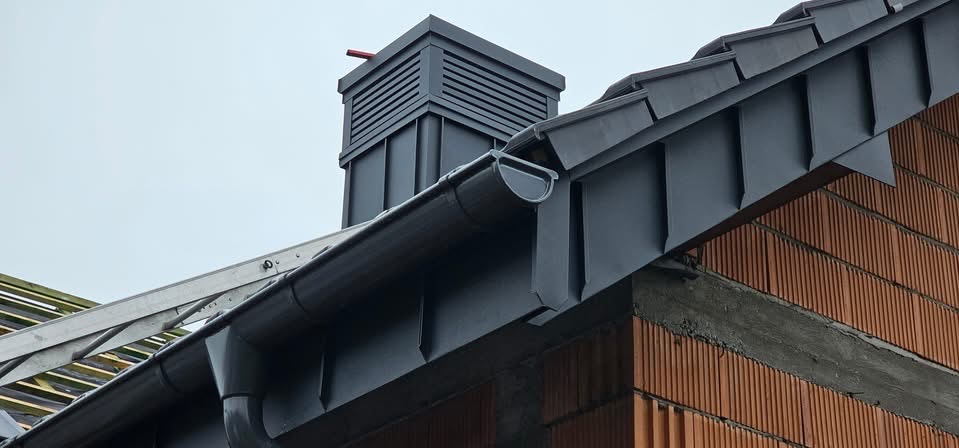 Szary system rynnowy i obróbki blacharskie na dachu z cegły, widok z dołu, fragment drabiny oparty o dach. Nowoczesny komin wentylacyjny.