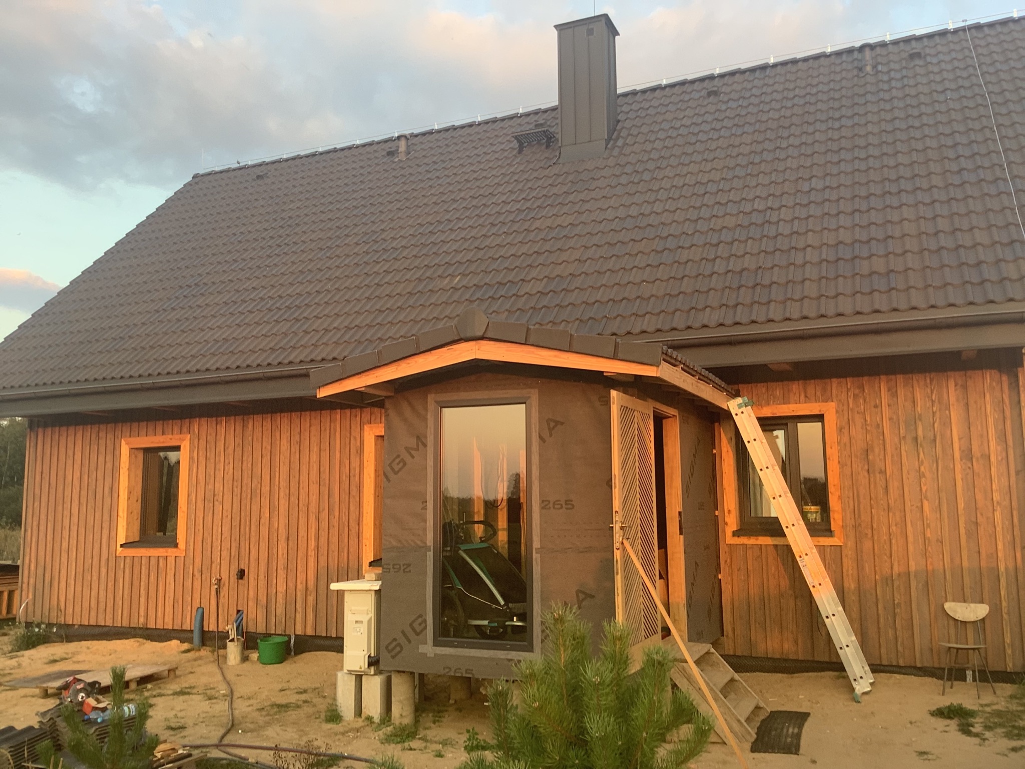 Dom z drewnianą elewacją i nowym, grafitowym dachem z kominem. Widoczna drabina oparta o ścianę, okna z czarną folią montażową. Prace wykończeniowe.