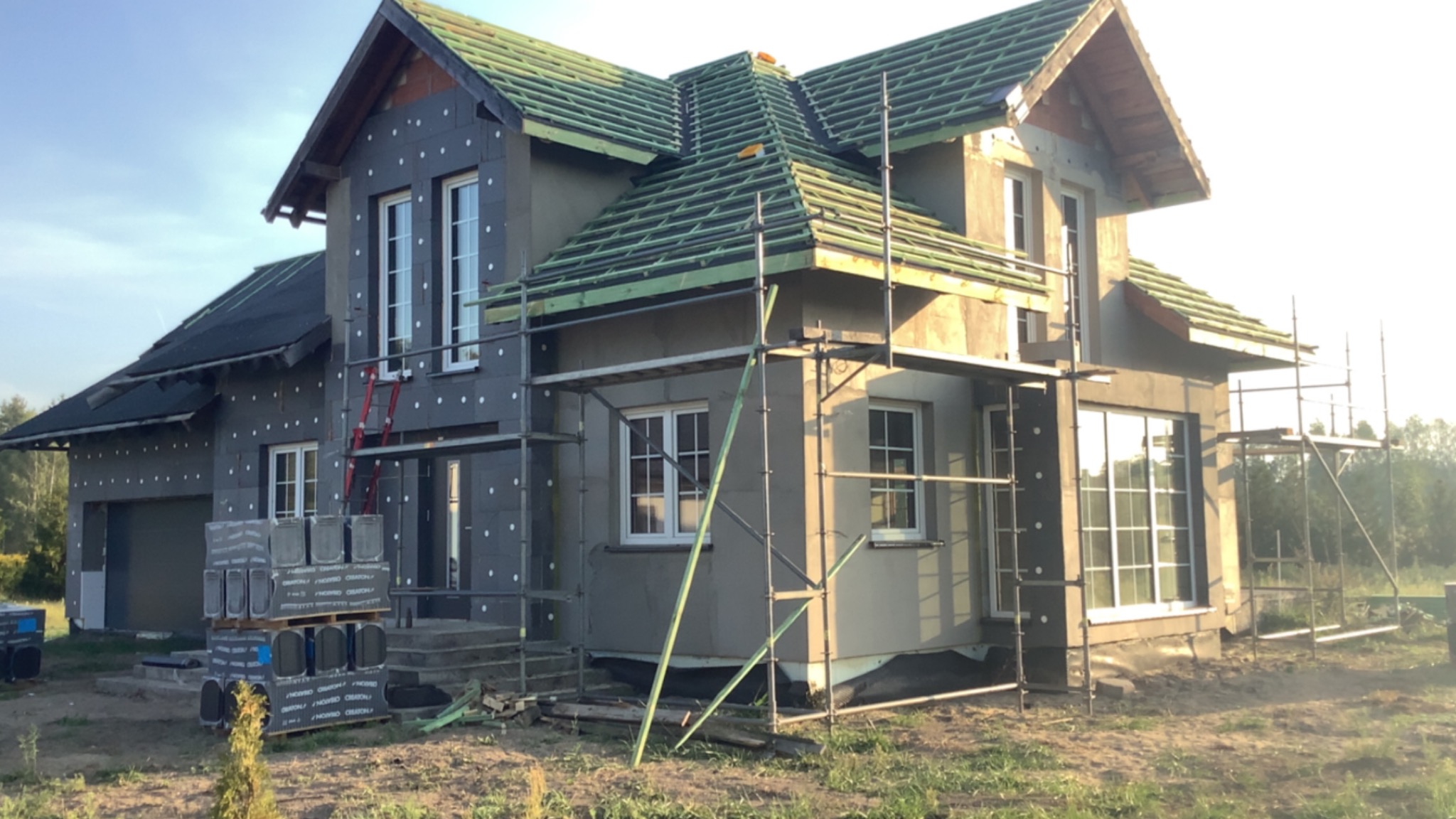 Dom w trakcie ocieplania styropianem, z widocznym rusztowaniem i częściowo wykończonym dachem. Na pierwszym planie paleta z materiałami budowlanymi.
