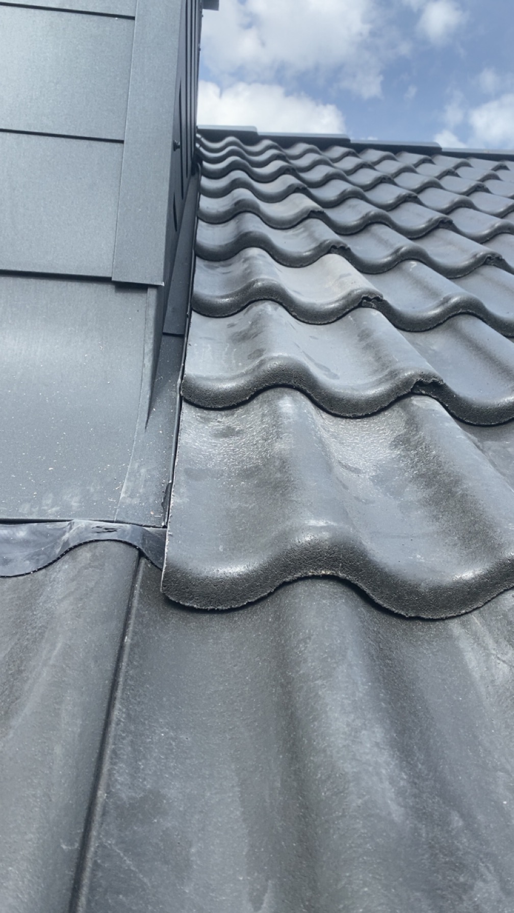 Szczegółowe ujęcie szarej, falistej dachówki ceramicznej, z widocznym połączeniem z obróbką blacharską i fragmentem elewacji. Widoczne łączenie dachu z kominem.