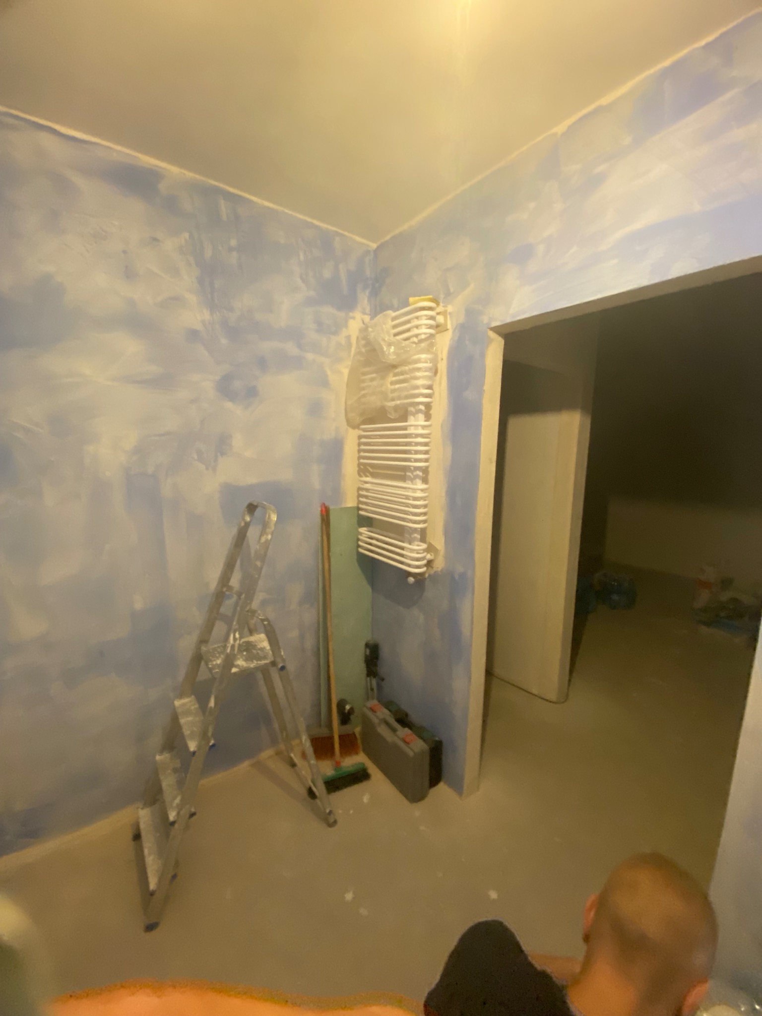 Pokój w trakcie malowania ścian na niebiesko, drabina, grzejnik owinięty folią, narzędzia malarskie. Widoczny fragment osoby w dolnej części kadru.