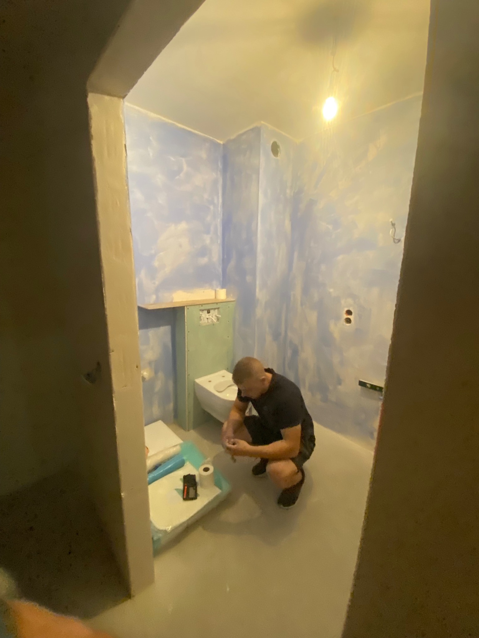 Remont łazienki: mężczyzna instaluje toaletę w pomieszczeniu z pomalowanymi na niebiesko ścianami. Widoczne narzędzia i materiały budowlane.