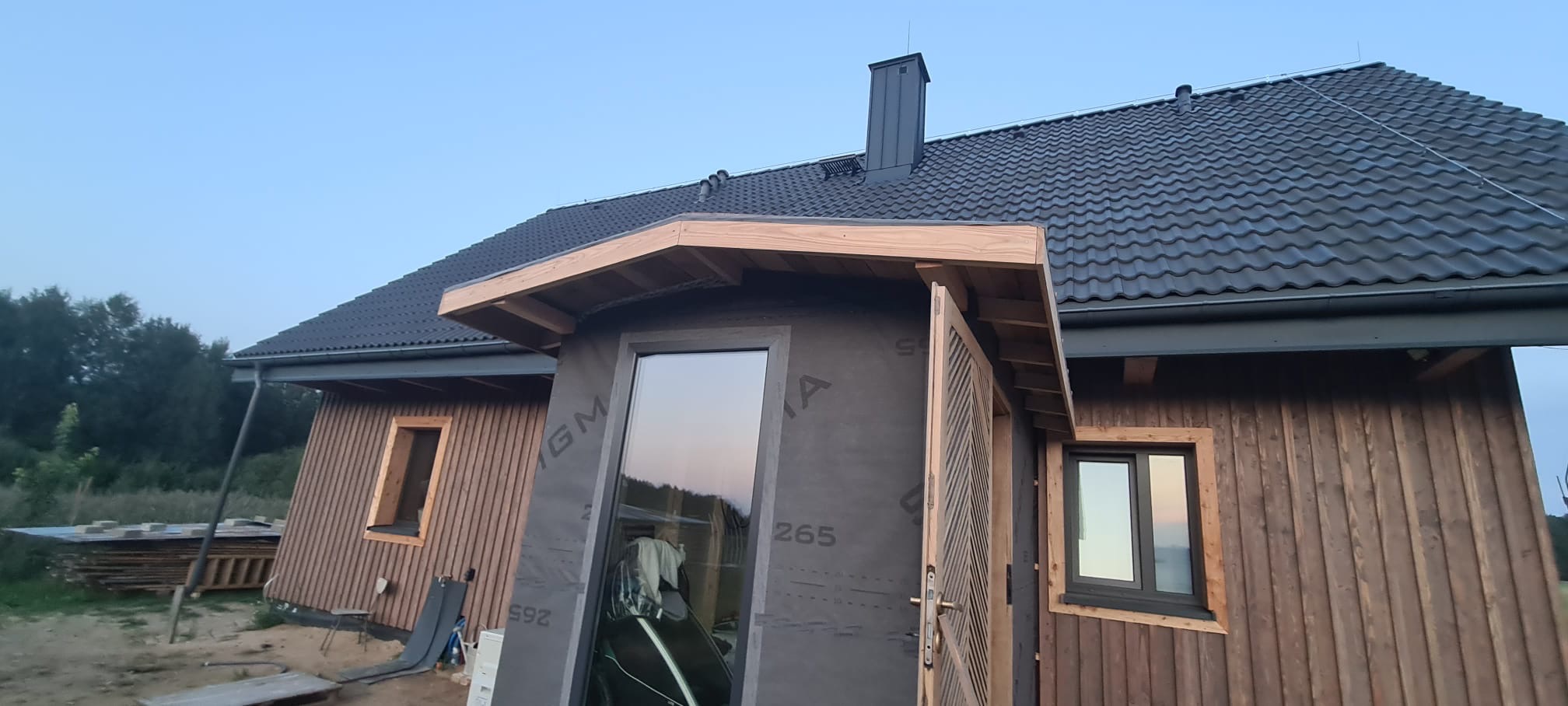 Dom w trakcie budowy z drewnianą elewacją, czarnym dachem i oknami. Widoczne detale wykończeniowe i materiały budowlane. Ujęcie z zewnątrz, wczesny etap prac.