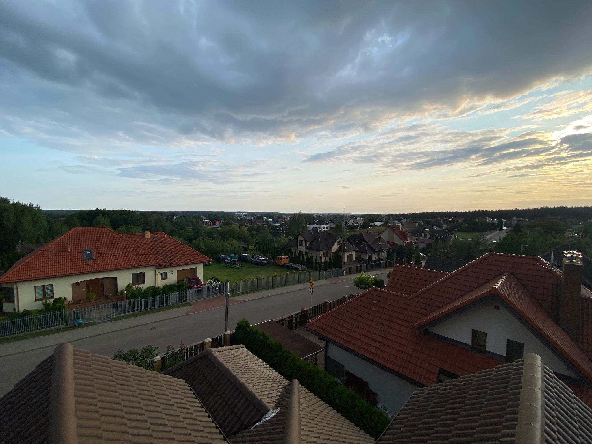 Panorama osiedla domów jednorodzinnych z perspektywy dachu, widoczne czerwone dachówki, ulice i zieleń, pochmurne niebo o zmierzchu.