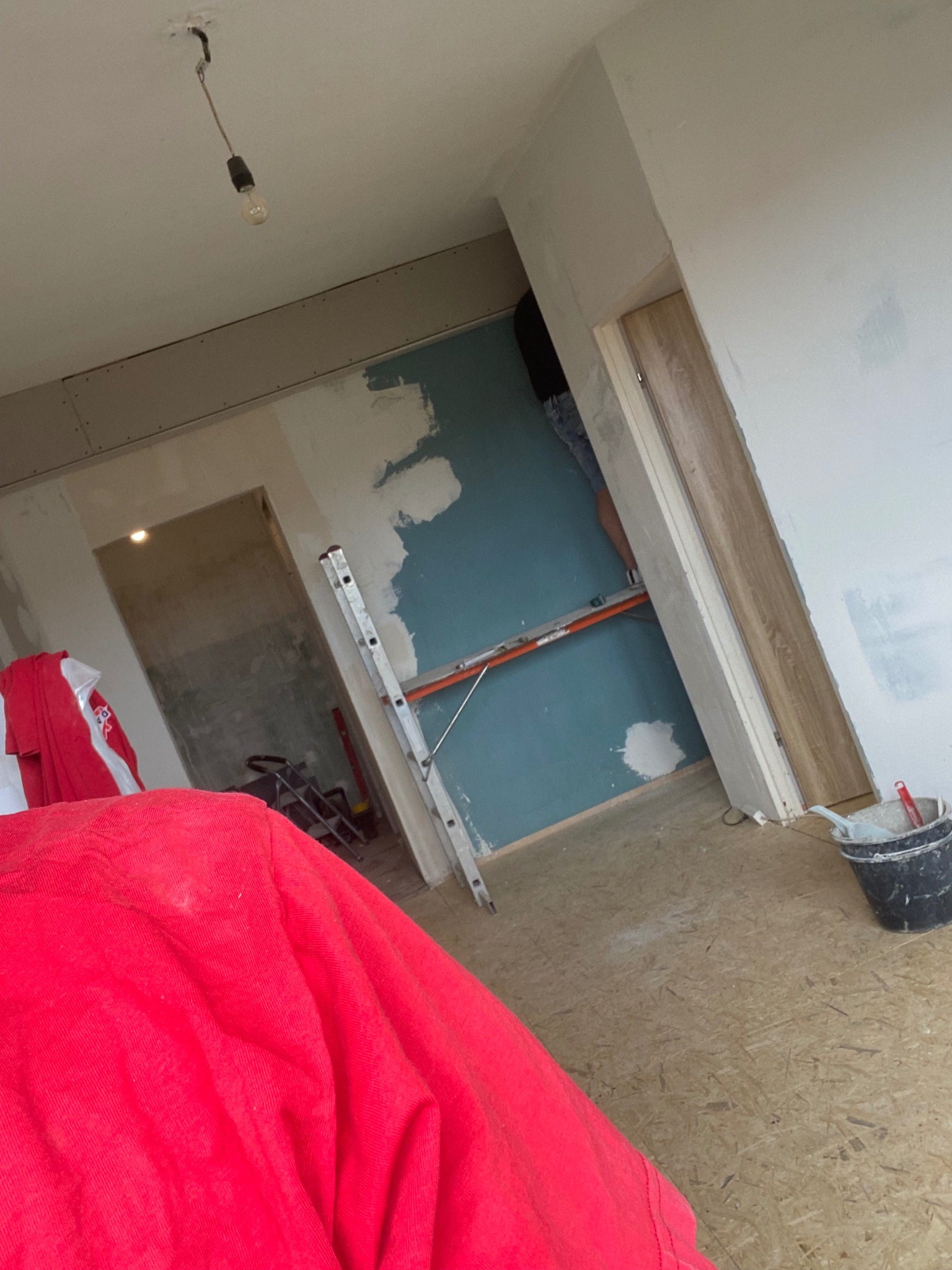 Wnętrze w trakcie remontu: ściana z częściowo nałożoną gładzią, drabina oparta o ścianę, widoczny fragment podłogi i wiadro z zaprawą. W tle widoczne kolejne pomieszczenie.