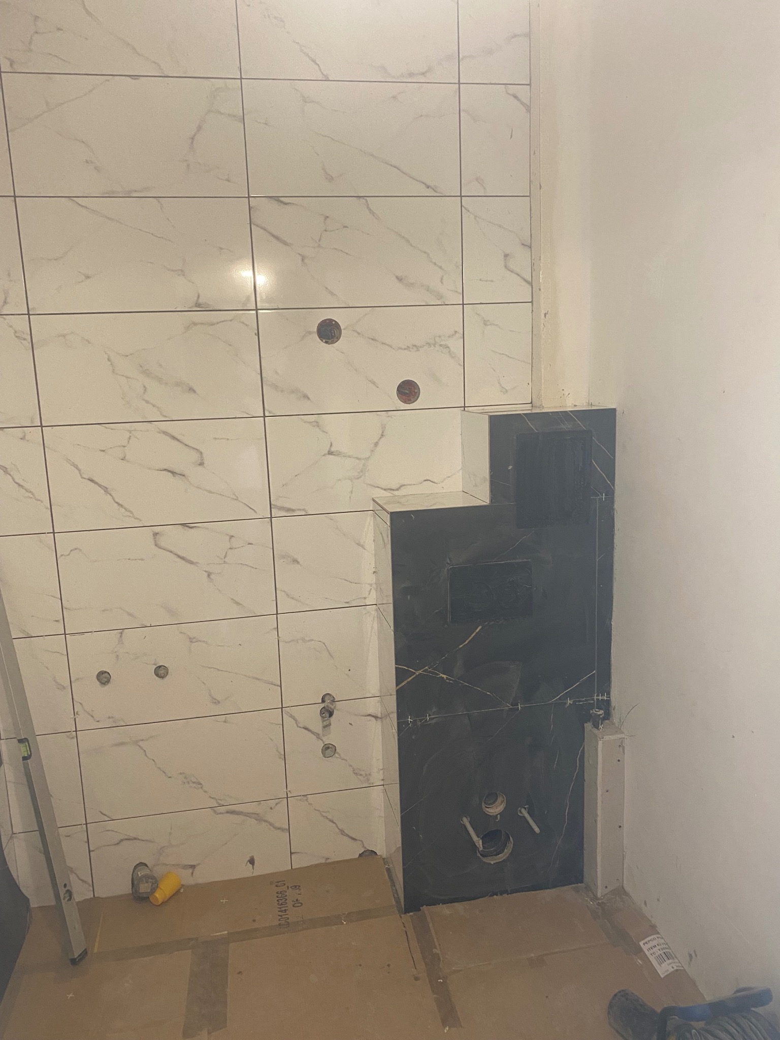 Wnętrze łazienki w trakcie remontu: białe płytki imitujące marmur na ścianach, czarne płytki wokół instalacji, widoczne otwory na armaturę, podłoga zabezpieczona kartonem.