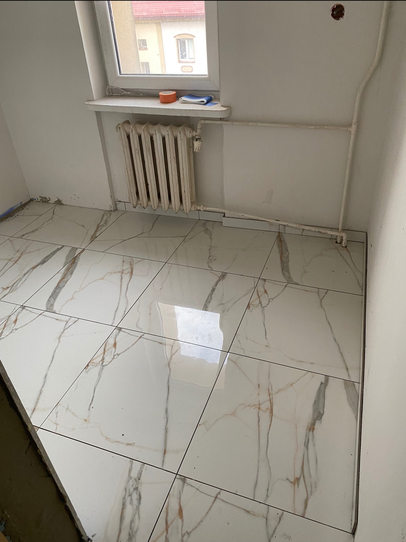 Położone białe płytki podłogowe z marmurowym wzorem w pokoju z grzejnikiem i oknem. Widoczne niedokończone wykończenia ścian.