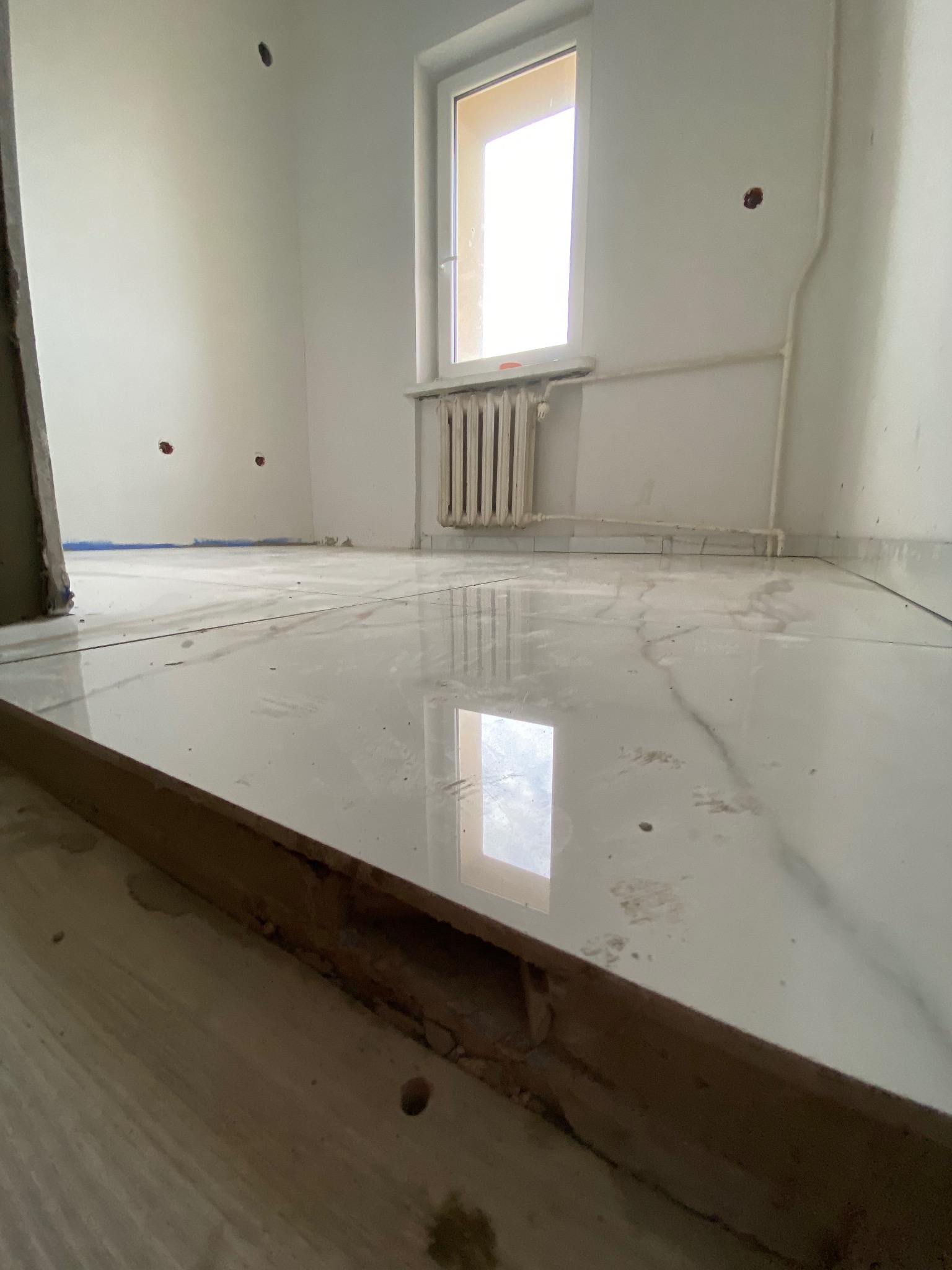 Błyszcząca, marmurowa podłoga po remoncie w jasnym pokoju z oknem i grzejnikiem. Widoczne ślady po wierceniu w ścianie. Ujęcie z niskiej perspektywy.