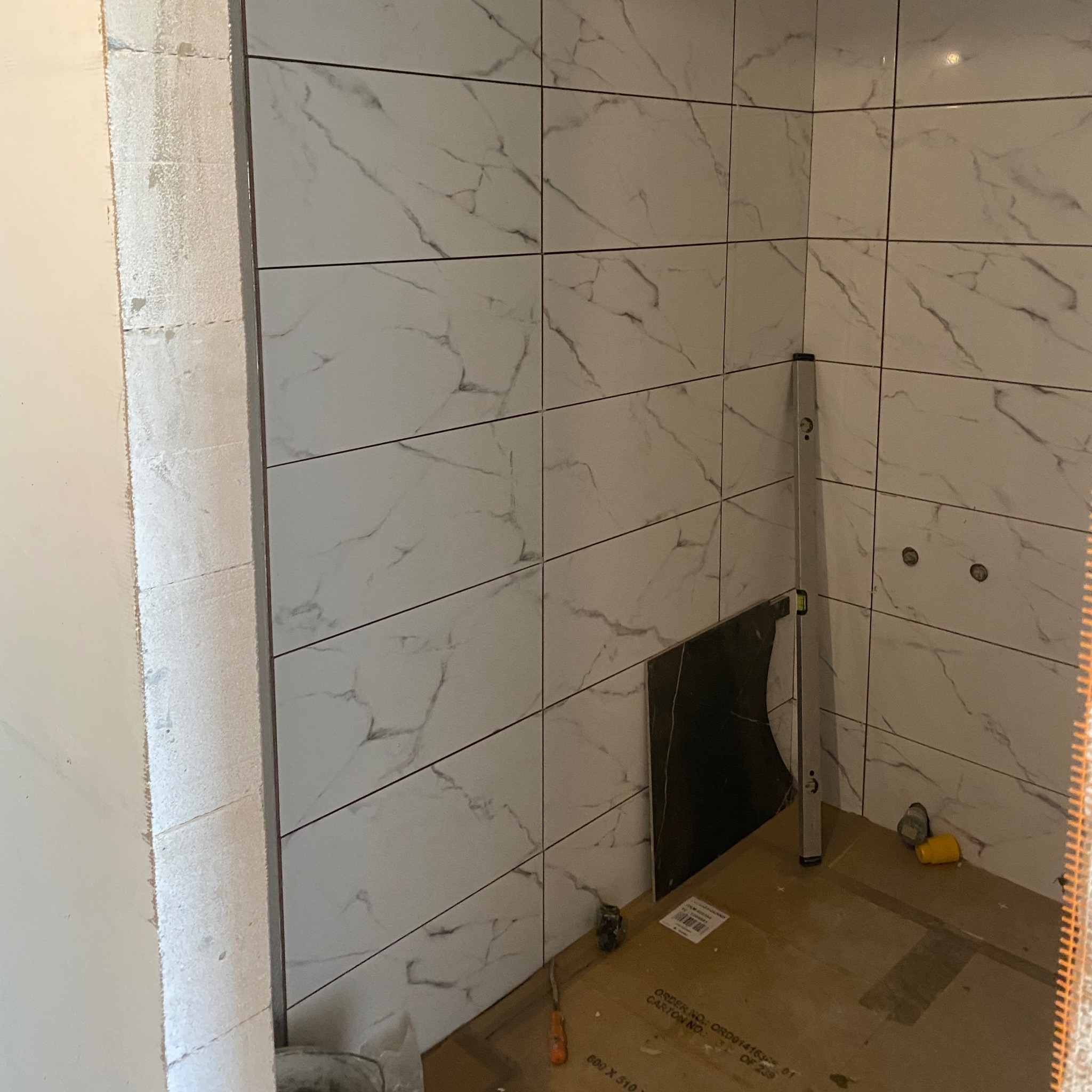 Układanie białych płytek imitujących marmur w rogu łazienki. Widoczna poziomica, fragment czarnej płytki i narzędzia na podłodze. Ściana przygotowana pod płytki.