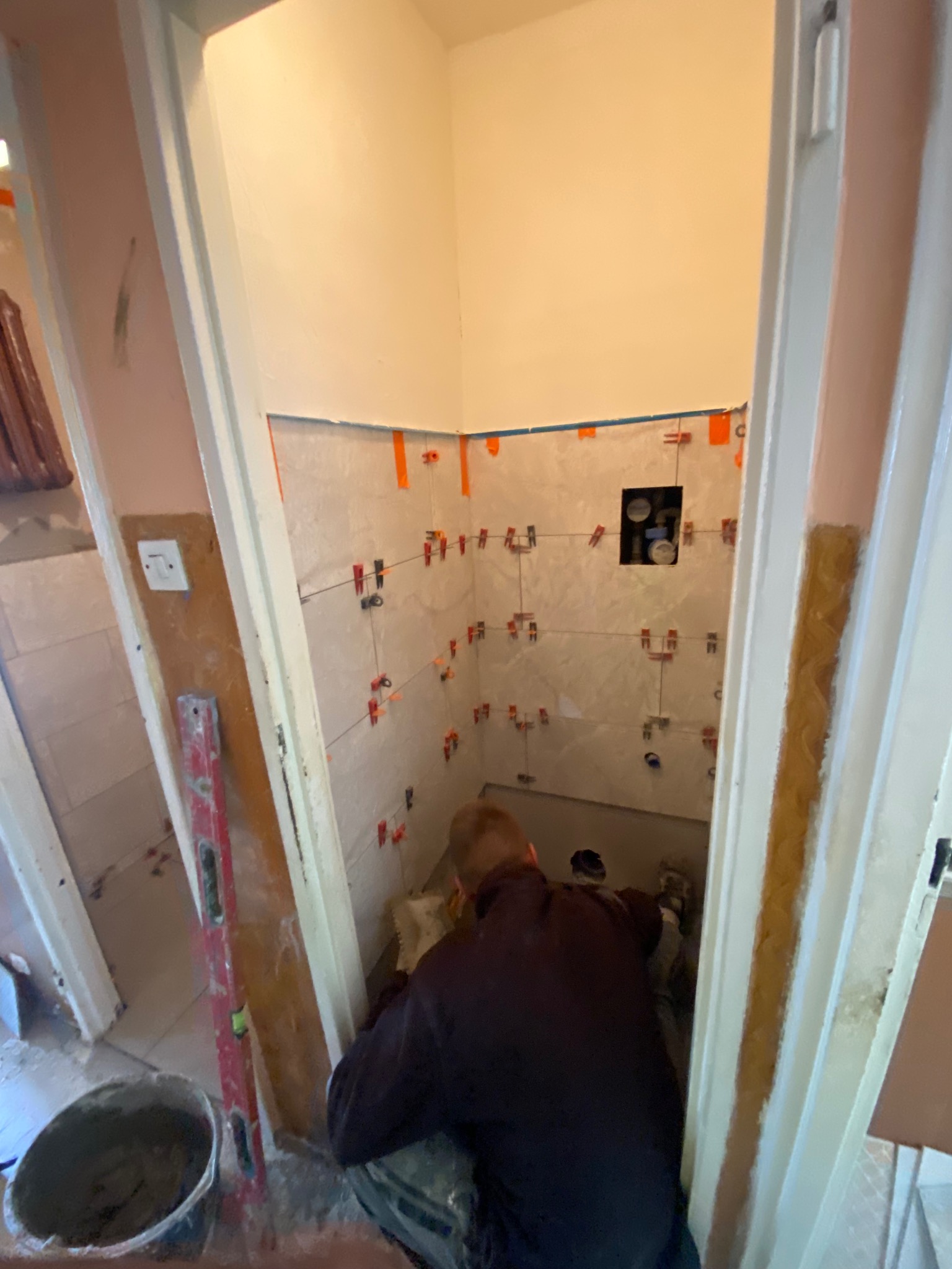 Układanie płytek w małej łazience z użyciem poziomicy i krzyżyków dystansowych. Widoczny fragment instalacji hydraulicznej. Praca w toku, wnętrze w trakcie remontu.