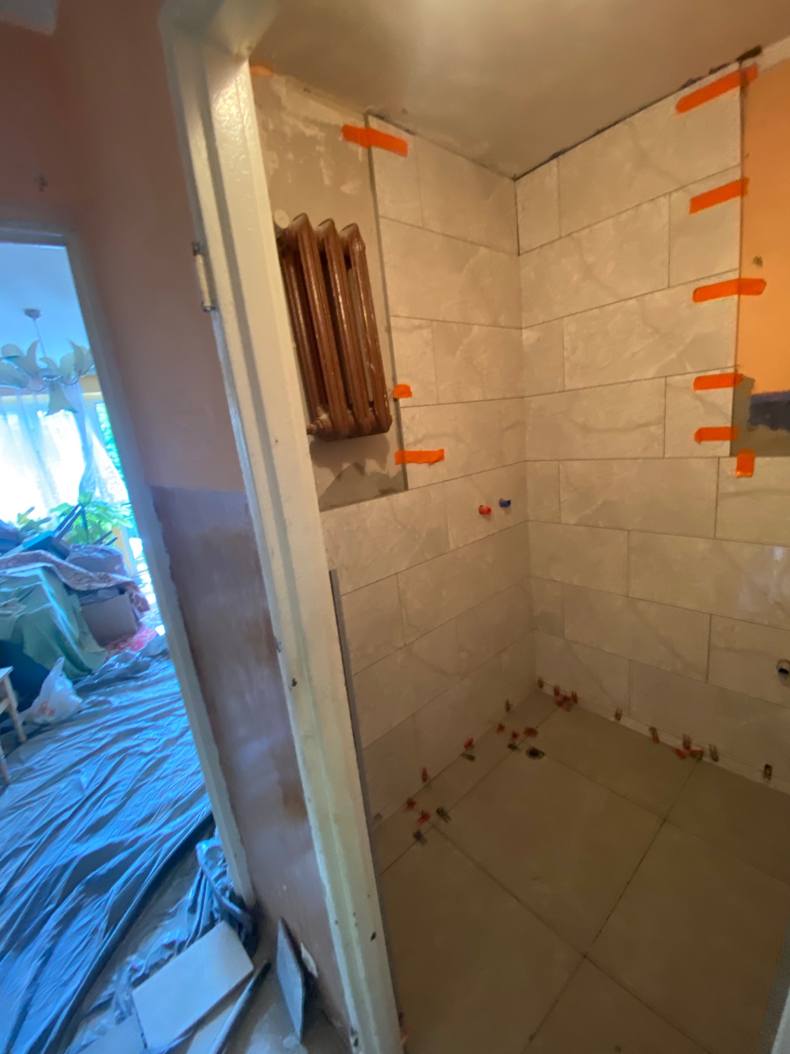 Wnętrze łazienki w trakcie remontu: ułożone jasne płytki na ścianach z pomarańczowymi krzyżykami dystansowymi, brązowy grzejnik na ścianie, podłoga w trakcie układania płytek.