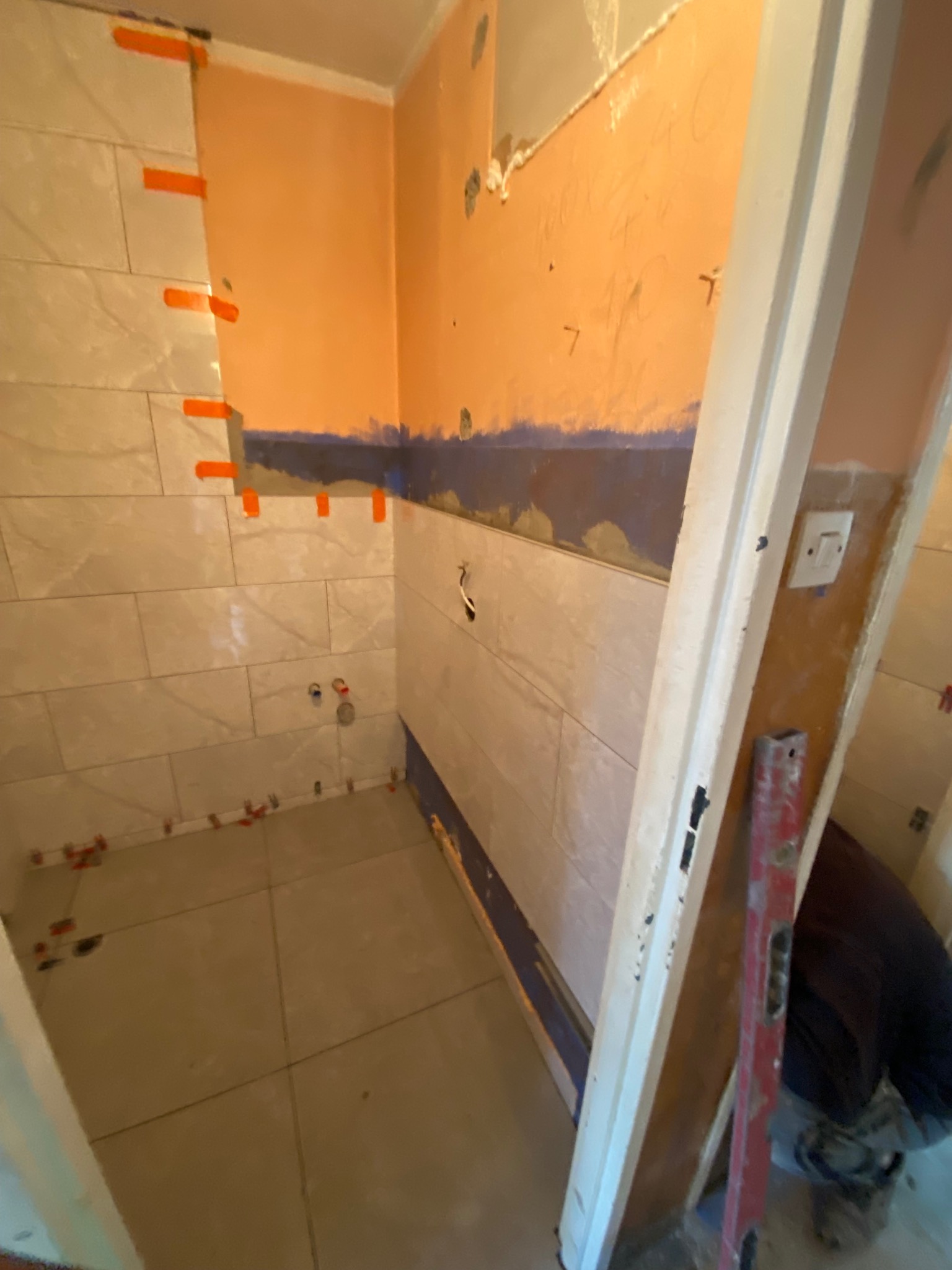 Wnętrze łazienki w trakcie remontu: częściowo ułożone płytki na ścianach i podłodze, poziomica oparta o futrynę, pomarańczowe taśmy i krzyżyki dystansowe.