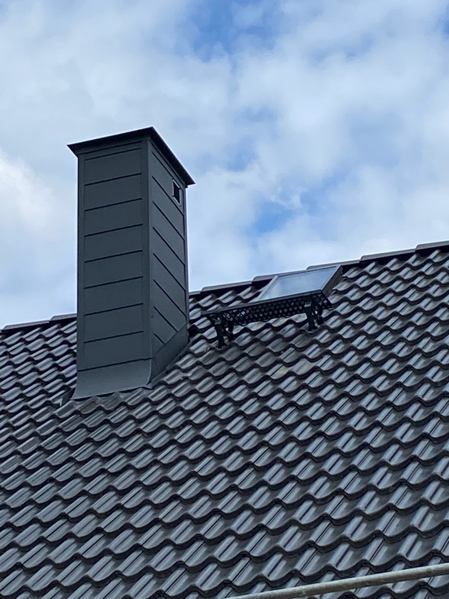 Grafitowy komin z blachy trapezowej i okno dachowe na ciemnej dachówce karpiówce, na tle błękitnego nieba z chmurami. Widok z dołu.