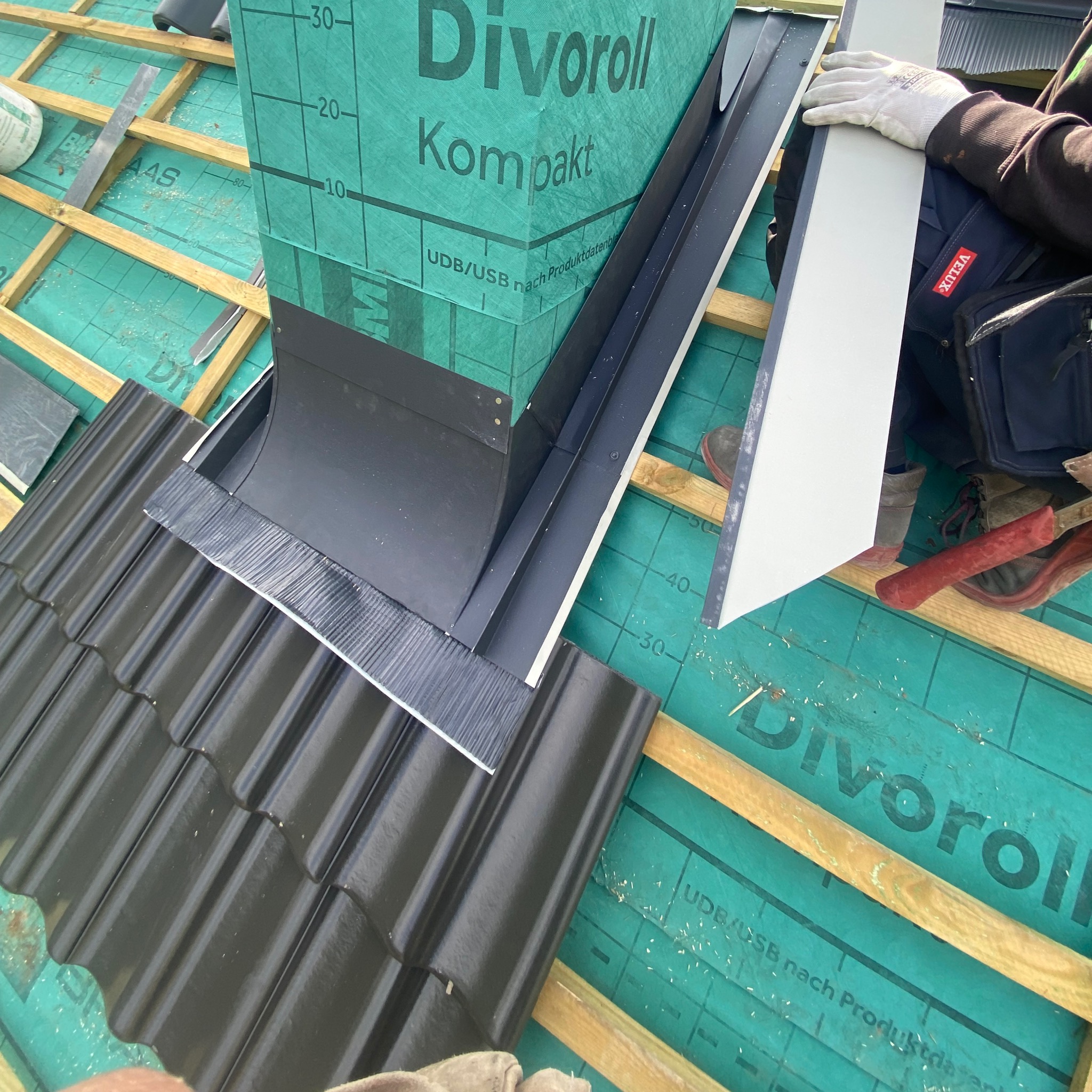 Montaż obróbki blacharskiej komina na dachu pokrytym czarną dachówką. Widoczna membrana dachowa Divoroll i narzędzia pracy dekarza. Praca w toku, zbliżenie.