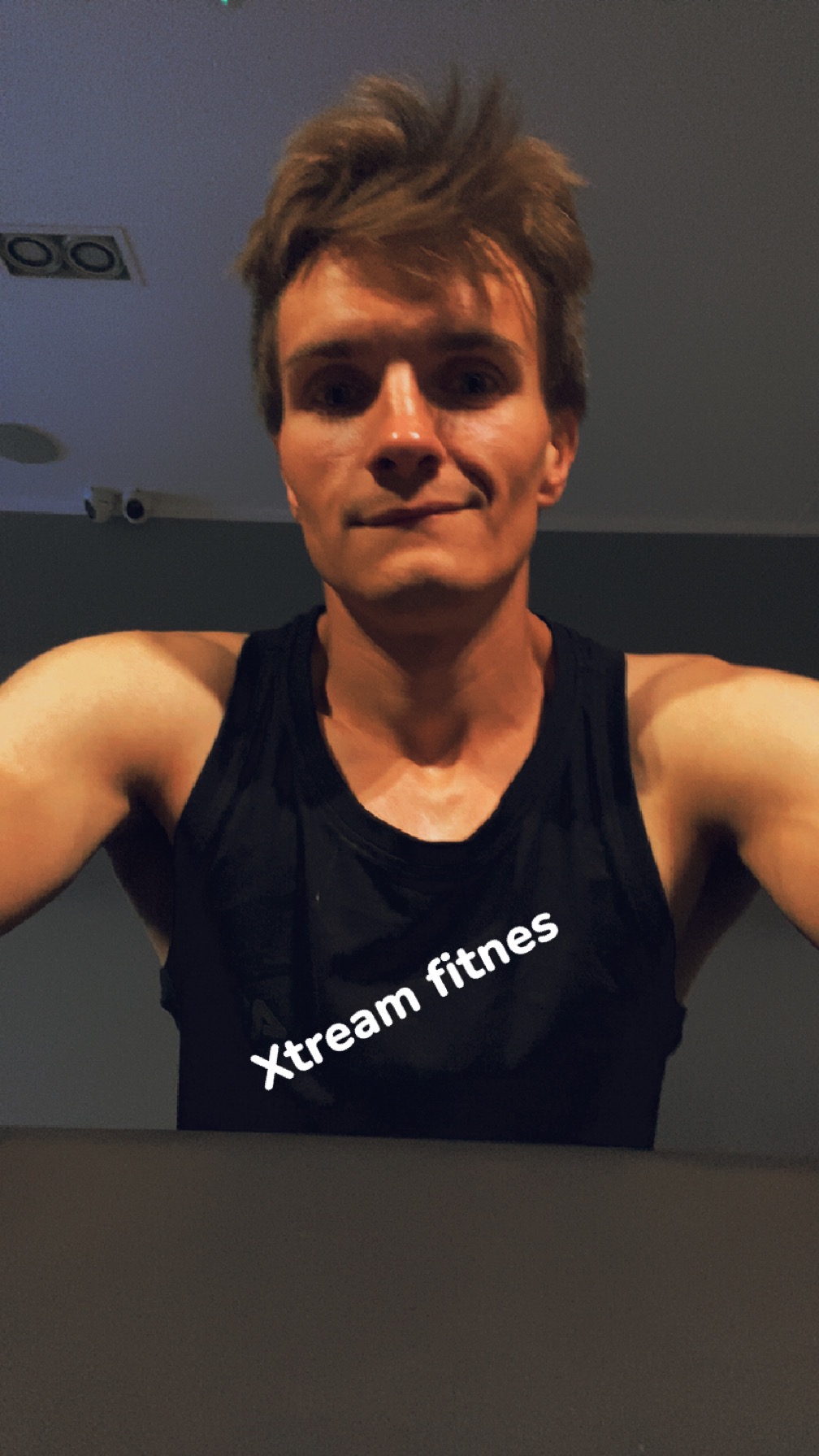 Młody mężczyzna w czarnej koszulce z napisem 'Xtream fitnes' pozuje do selfie w siłowni. Widoczne kamery monitoringu na suficie. Ujęcie z dołu.