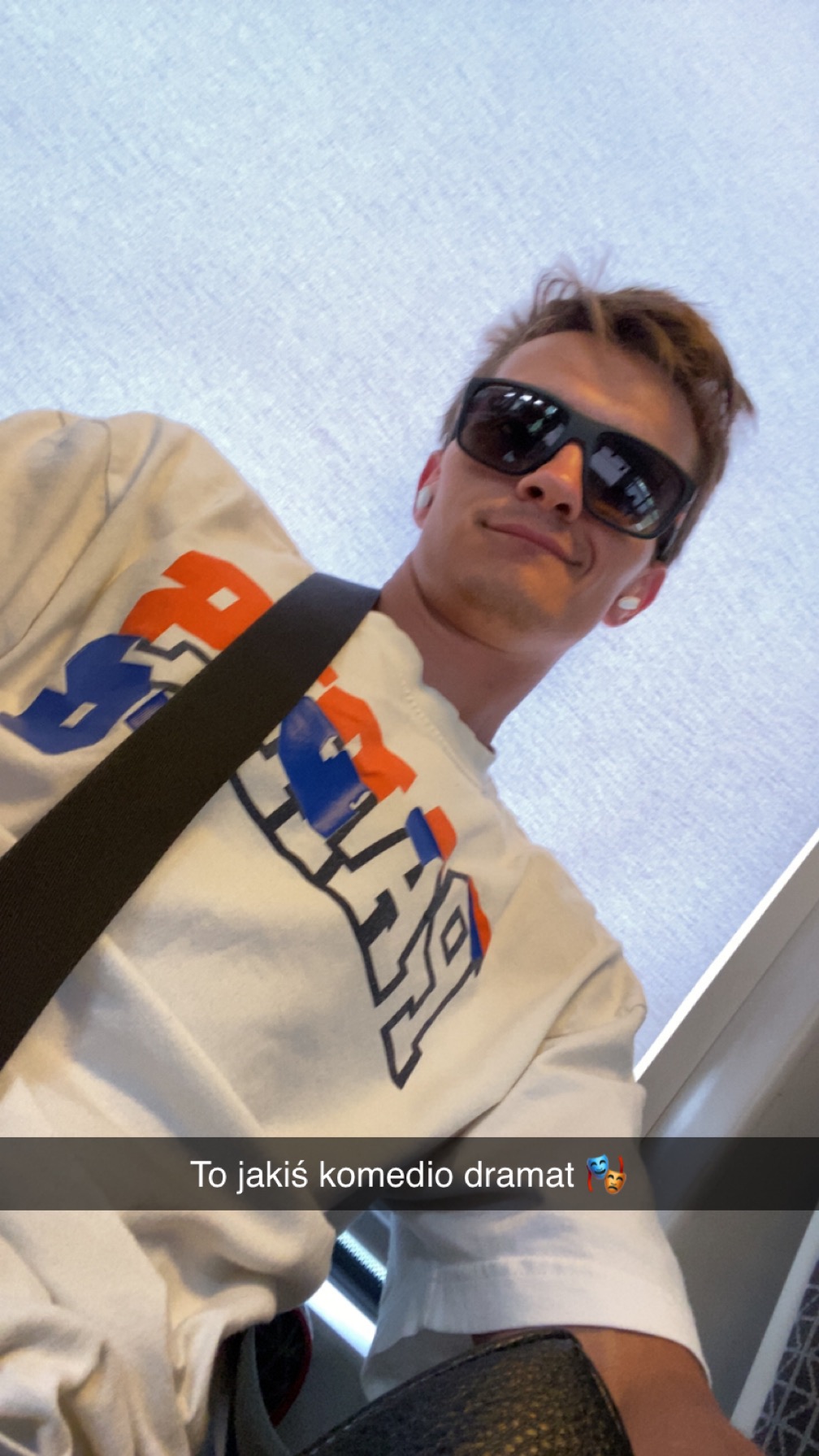 Mężczyzna w okularach przeciwsłonecznych i białej koszulce z nadrukiem, robiący selfie pod nietypowym kątem, z widocznym paskiem i fragmentem tekstu.