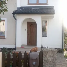 Dwukondygnacyjny dom z ciemną dachówką i rynnami, kremową elewacją i brązową stolarką okienną. Widoczny fragment ogrodzenia z skrzynką na listy.